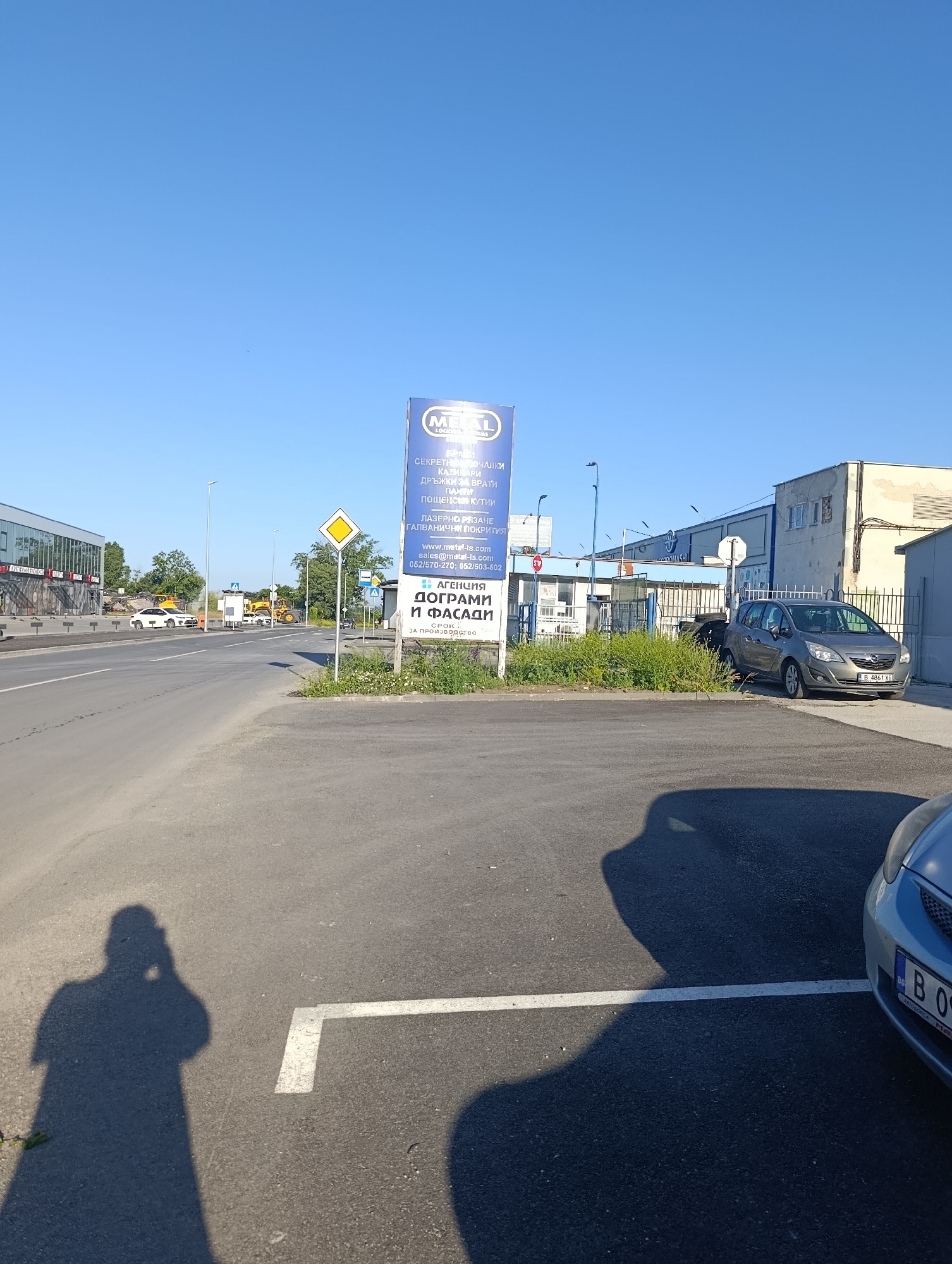 The image depicts an outdoor scene on a clear day, likely in an industrial or commercial area. A tall blue and white sign stands prominently on the right side of the frame, advertising "METAL LS" and listing services such as "СЕКРЕТНИ КОМПЛЕКТИ," "КАТИ," "АРМИ," "ДРЪЖКИ ЗА ВРАТИ," "ТОЧКИ," and "ПОЦЕНСКИ КУТИИ." Below these services, it lists "ЛАЗЕРНО РЯЗАНЕ," "ГАЛВАНИЧНИ ПОКРИТИЯ," along with contact information including a website and phone numbers.
Further down the sign, it indicates "АГЕНЦИЯ," "ДОГРАМИ И ФАСАДИ," and "СРОК / ЗА ПРОИЗВОДСТВО." In front of the sign, there is overgrown green grass and some weeds.
To the right of the sign, a grey hatchback car is parked on a paved surface, with its license plate visible as "B 4861 XE." Behind the car is a fenced area with some industrial buildings. The buildings appear to be made of concrete and metal, with some windows and corrugated metal siding. One building has "MOTOWASH" written on its facade.
In the foreground, a large shadow of a person holding a phone is cast on the asphalt parking lot. To the right, the front of a blue car is partially visible, with its license plate obscured by the shadow.
The background shows a road with cars, including a white sedan and construction vehicles like yellow excavators. There are streetlights and power lines visible. The sky is clear and blue. The overall environment suggests a business district or an area with industrial activity.