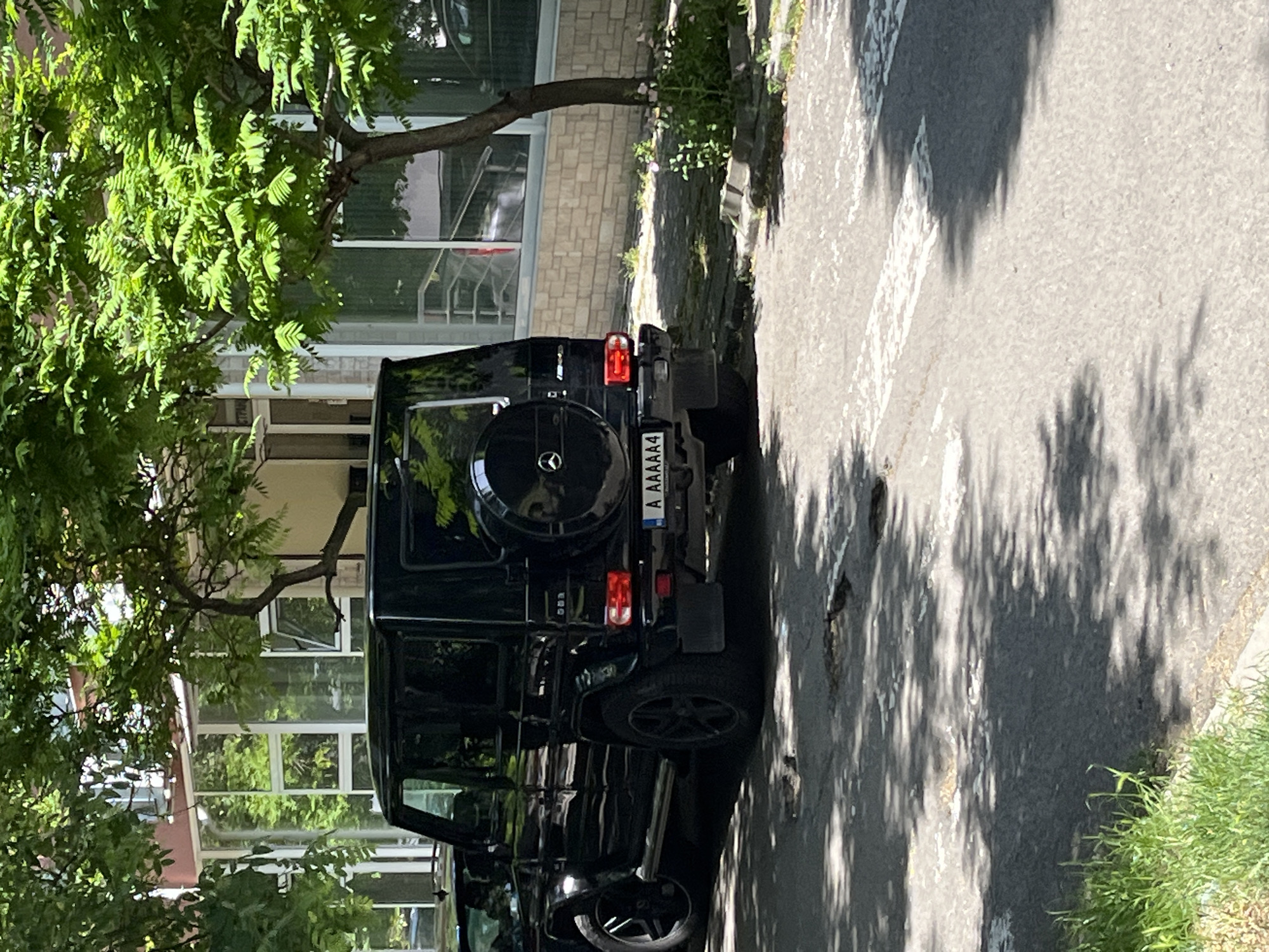 The image displays a black Mercedes-Benz G-Class (G-Wagen) parked outdoors, viewed from its rear passenger side. The vehicle is situated on a paved surface, likely an asphalt street or parking area, with strong, dappled shadows from overhead trees indicating bright sunshine.
Lush green tree foliage dominates the left side of the frame, partially obscuring a multi-story building behind it. The building features a light-colored, possibly brick or stone facade on its lower part and numerous large windows above, reflecting elements of the surroundings. A narrow strip of green grass and some sparse vegetation runs along the edge of the paved surface next to the building.
The rear of the G-Wagen clearly shows its spare tire, mounted on the tailgate and featuring the Mercedes-Benz logo. Below the rear window, the license plate is visible and reads "A AAAAA4". The vehicle's tail lights, bumper, and parts of its exhaust system are also discernible. The overall scene suggests a sunny daytime setting.