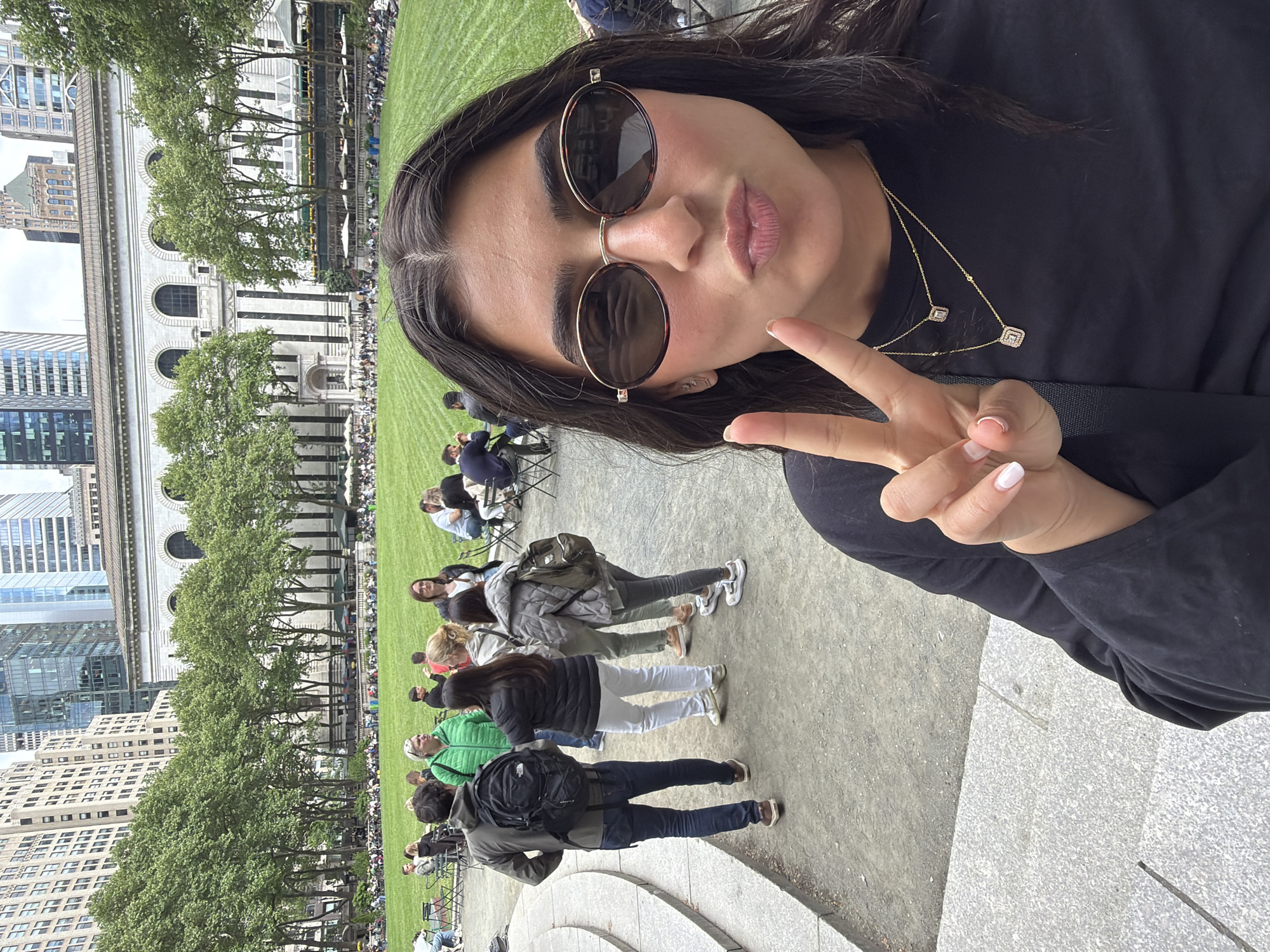 A woman is shown in a selfie-style image at Bryant Park in New York City. The park is populated with people relaxing on the lawn and in chairs. There are trees and tall buildings in the background. The woman is wearing sunglasses, a black long-sleeved shirt, and a gold necklace. She is making a peace sign with her hand and pursing her lips.