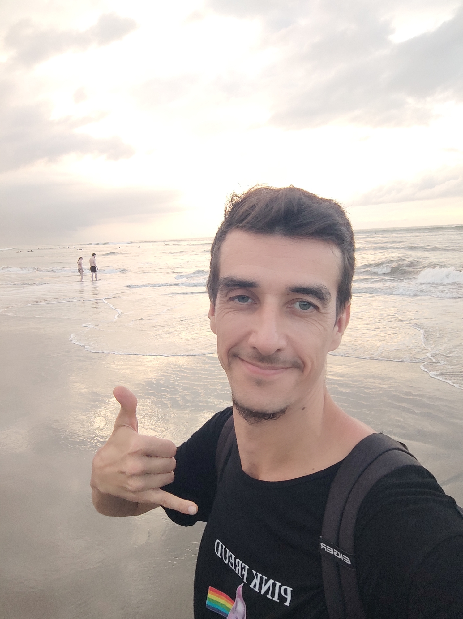 A man takes a selfie on a beach in North Kuta, Indonesia. He has dark hair, a subtle goatee, and light eyes, and is smiling pleasantly at the camera. He is making a "shaka" hand gesture with his left hand, extending his thumb and pinky finger. He wears a black t-shirt with a reversed graphic featuring the text "PINK FLOYD" above a rainbow prism. A dark backpack, with the reversed brand name "EIGER" visible on a strap, rests on his shoulders.
The setting is a tranquil beach scene, likely during either dawn or dusk, given the soft, diffused sunlight filtering through an overcast sky. The sky is bright and hazy with visible cloud formations. The ocean's gentle waves lap onto the shore, creating wet, reflective sand that mirrors the soft light from the sky. In the mid-ground, two people are standing in the shallow surf, while further out in the water, several small figures, possibly surfers, are visible on the horizon. The overall atmosphere conveyed is peaceful and relaxed.