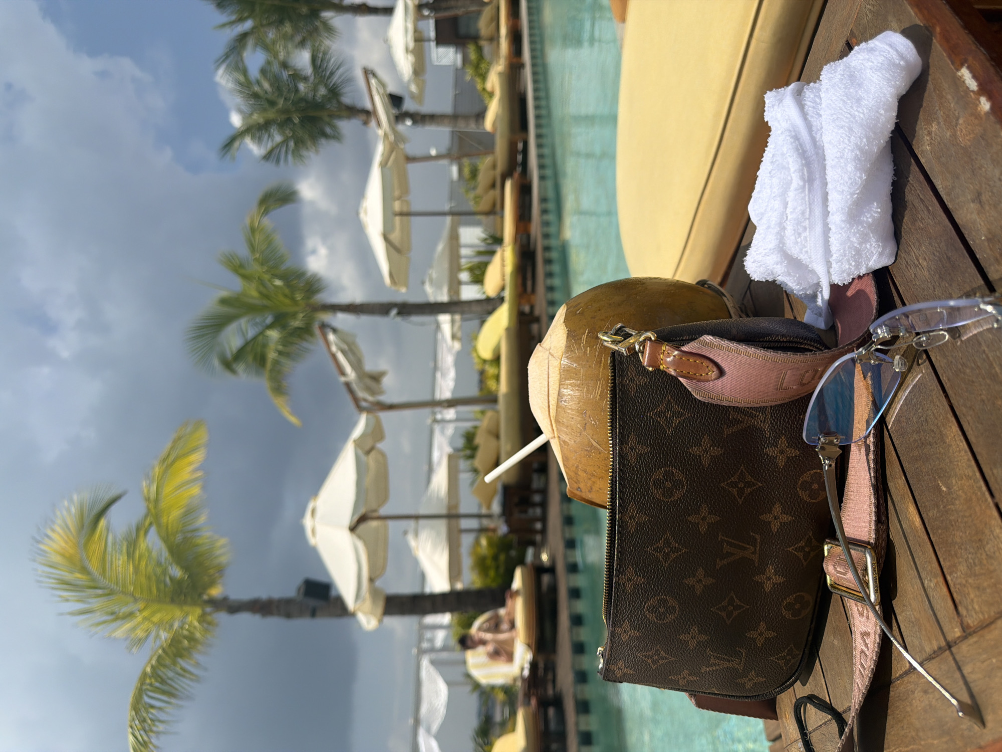The scene depicts a poolside area in Tibubeneng, Indonesia, viewed from a low perspective. In the immediate foreground, on a weathered brown wooden surface, are a brown Louis Vuitton monogram canvas pouch with a pink strap, a whole coconut with a white straw inserted, a pair of rimless sunglasses with blue-tinted lenses, a small folded white terry cloth towel, and a black elastic hair tie.
Beyond these objects, a clear blue swimming pool extends across the midground. Along the far edge of the pool, multiple yellow sun loungers are arranged, each with a white parasol. One individual is seated on a yellow and white striped sun lounger in the left midground, facing away from the camera, and appears to be interacting with a small object held in their hands.
The background features several tall palm trees with green fronds, interspersed among additional white parasols and yellow sun loungers. The sky above is partially overcast, with dark grey clouds dominating the upper left portion, transitioning to a lighter blue sky with scattered white clouds on the right. The environment suggests a resort or leisure facility.