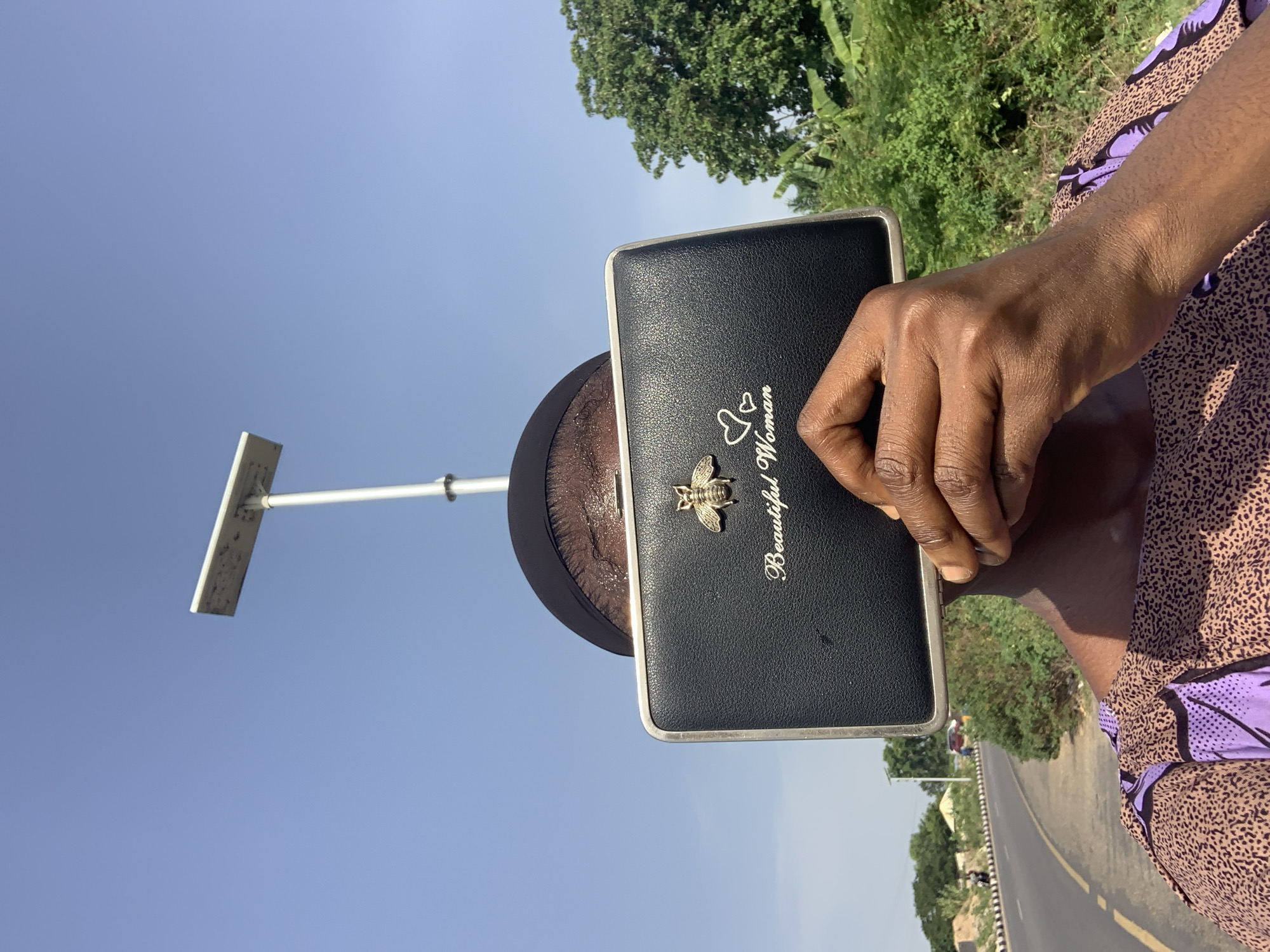 A person holds a black clutch that says "Beautiful Woman" with a bee charm, outdoors on a sunny day near a road in Zango-ndaji, Nigeria. The person's hand is visible and they are wearing a patterned shirt. A street light pole is visible in the background, along with trees and a road.