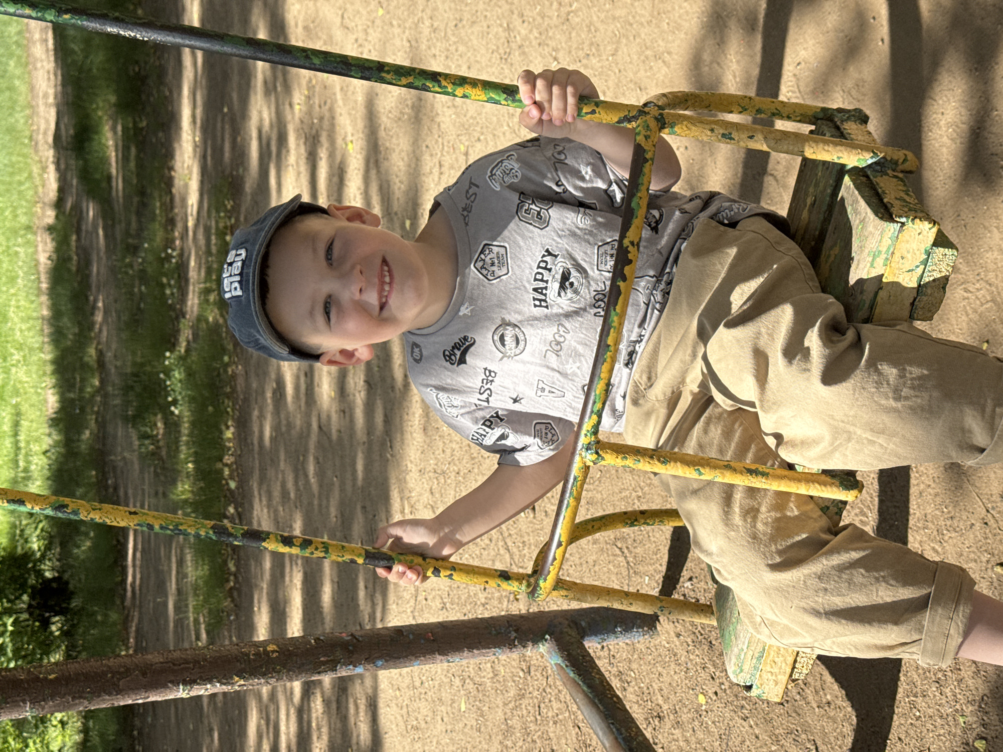 A young boy wearing a grey t-shirt with graphics and a dark baseball cap is sitting on a yellow and green swing. He is smiling directly at the camera. The swing is made of wood and metal, and it appears to be in a park or playground. The ground around the swing is sandy or dirt. In the background, there are trees and green grass, suggesting an outdoor, natural environment. The image is taken in daylight, with shadows cast on the ground, indicating the sun is shining. The location is identified as Chaikovsky, Russia.