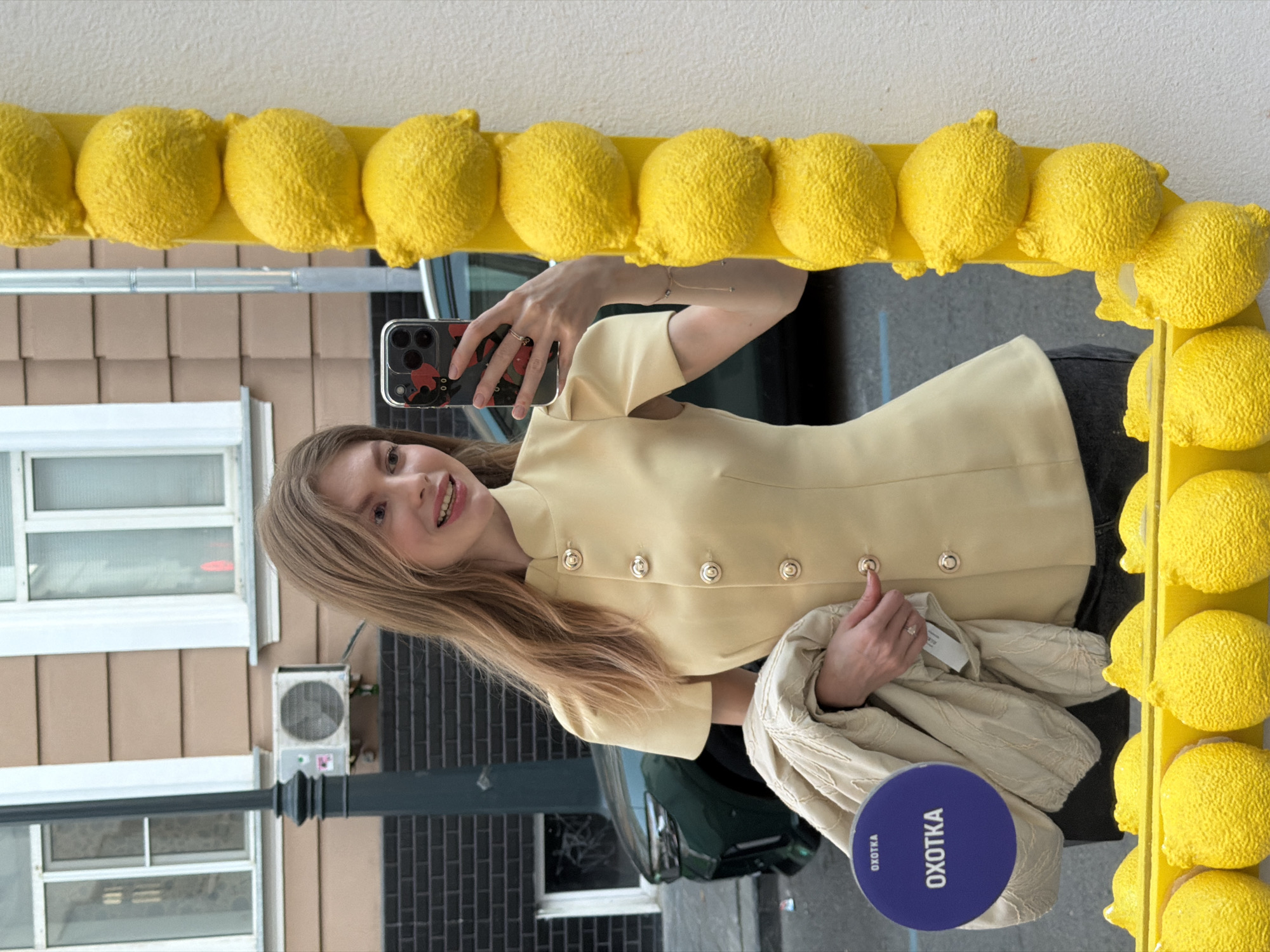 A young woman is taking a selfie in a mirror framed with yellow lemon decorations. She is holding a smartphone in her right hand. The mirror is positioned outdoors, reflecting the city street behind her. The woman is wearing a light yellow jacket with silver buttons and dark pants. She is also holding a beige patterned garment in her left hand, topped with a round, navy blue object with the text "OXOTKA" printed in white.
The reflection in the mirror reveals a typical urban scene, including an air conditioning unit mounted on a wall, windows, a black lamppost, a brick wall, and a car. The city is Moscow, Russia, as indicated by the inscription "OXOTKA".