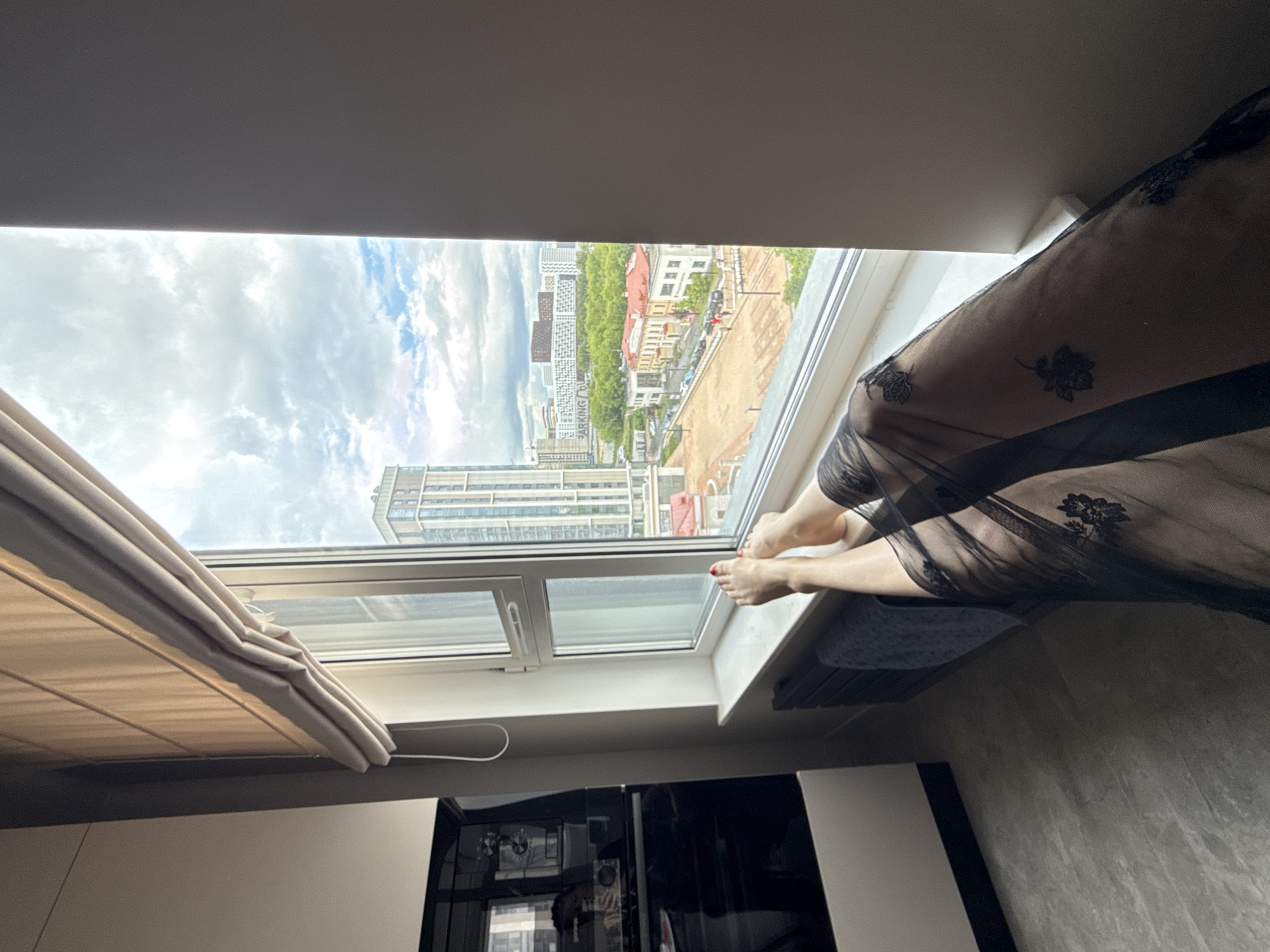 A medium-angle interior shot captures a view from a window in Dolgoprudny, Russia. A person sits on the windowsill, with their feet visible, draped in a sheer black robe adorned with floral embroidery. The window offers a view of a cityscape, featuring modern buildings and older, smaller structures. The sky is overcast with a mix of clouds. The bottom portion of the image includes elements of the interior, with a modern cabinet partially visible, and folded blinds covering a portion of the window.