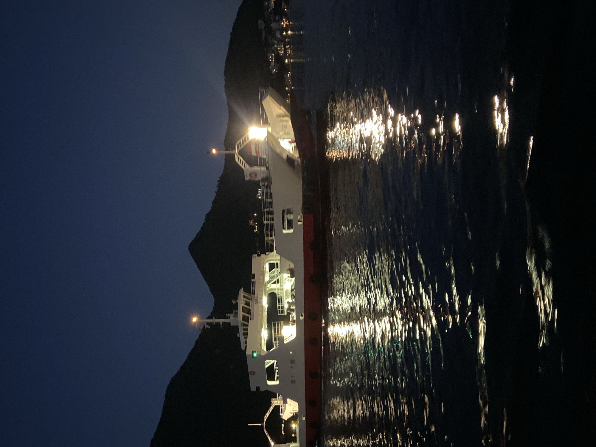 A ferry, identified by the name "GREBALI" on its side, is illuminated at night on a body of water, likely a bay or strait, near the town of Đurići, Montenegro. The ferry has a white upper structure with red trim along its waterline. Lights are visible on the ferry's decks and on a mast. The water reflects the ferry's lights, creating a shimmering effect. In the background, dark, silhouetted hills or mountains rise, with scattered lights indicating a settlement on the shore. The sky is dark, suggesting nighttime. There are no people clearly visible.