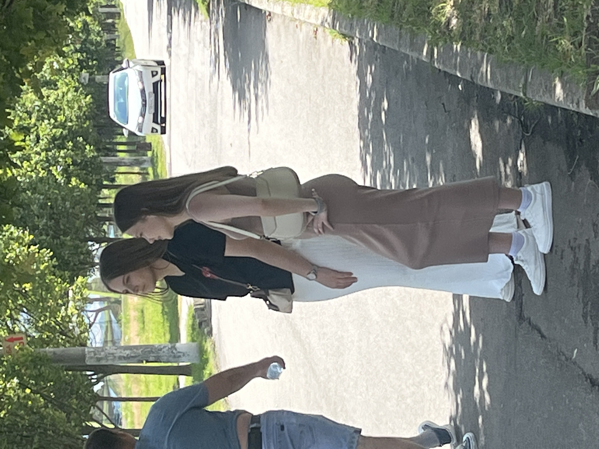The image depicts a street scene in Krasnodar, Russia, on a bright, sunny day. The orientation of the image is sideways. In the foreground, a person wearing a blue t-shirt and denim shorts is partially visible, holding a clear plastic bottle. Behind them, two young women are standing side-by-side, facing away from the camera and towards a road. The woman on the left is wearing a black t-shirt and a white skirt, and the woman on the right is wearing a beige top and a brown skirt. Both women have long, dark hair and are wearing white sneakers. The woman on the right has a small beige purse slung over her shoulder. The street is paved, with a concrete curb and some vegetation bordering it. In the background, a white car is parked on the side of the road, and trees line the area. The lighting suggests it is daytime, likely late morning or early afternoon, with strong shadows cast by the trees. There are no visible emotions conveyed by the subjects as they are not facing the camera. No text is clearly discernible in the image.
