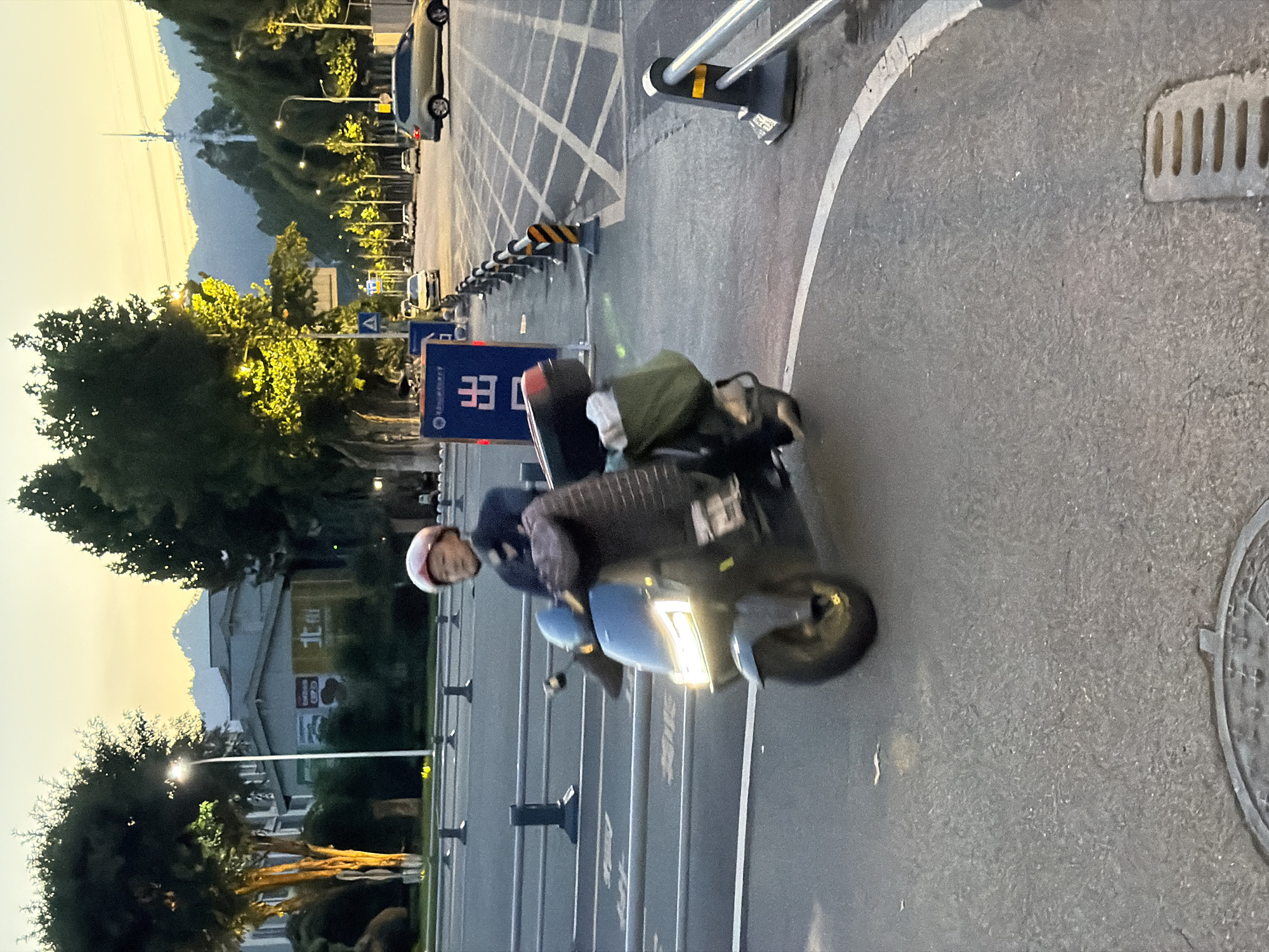 A person wearing a pink helmet and a dark jacket is riding a dark-colored electric scooter on a paved road at dusk. The scooter has a white illuminated headlight and a black storage box on the back, from which a green bag is hanging. The person is looking forward. The road appears to be in an urban area, with buildings and trees lining the side. In the background, mountains are visible against a hazy sky. There are other vehicles on the road, including cars and what appears to be another scooter. Traffic barriers and streetlights are also present. A sign with Chinese characters is visible on the side of the road. The scene appears to be in the city of Jiangjin, China, as indicated by the text on the sign. The overall lighting suggests it is either sunrise or sunset.