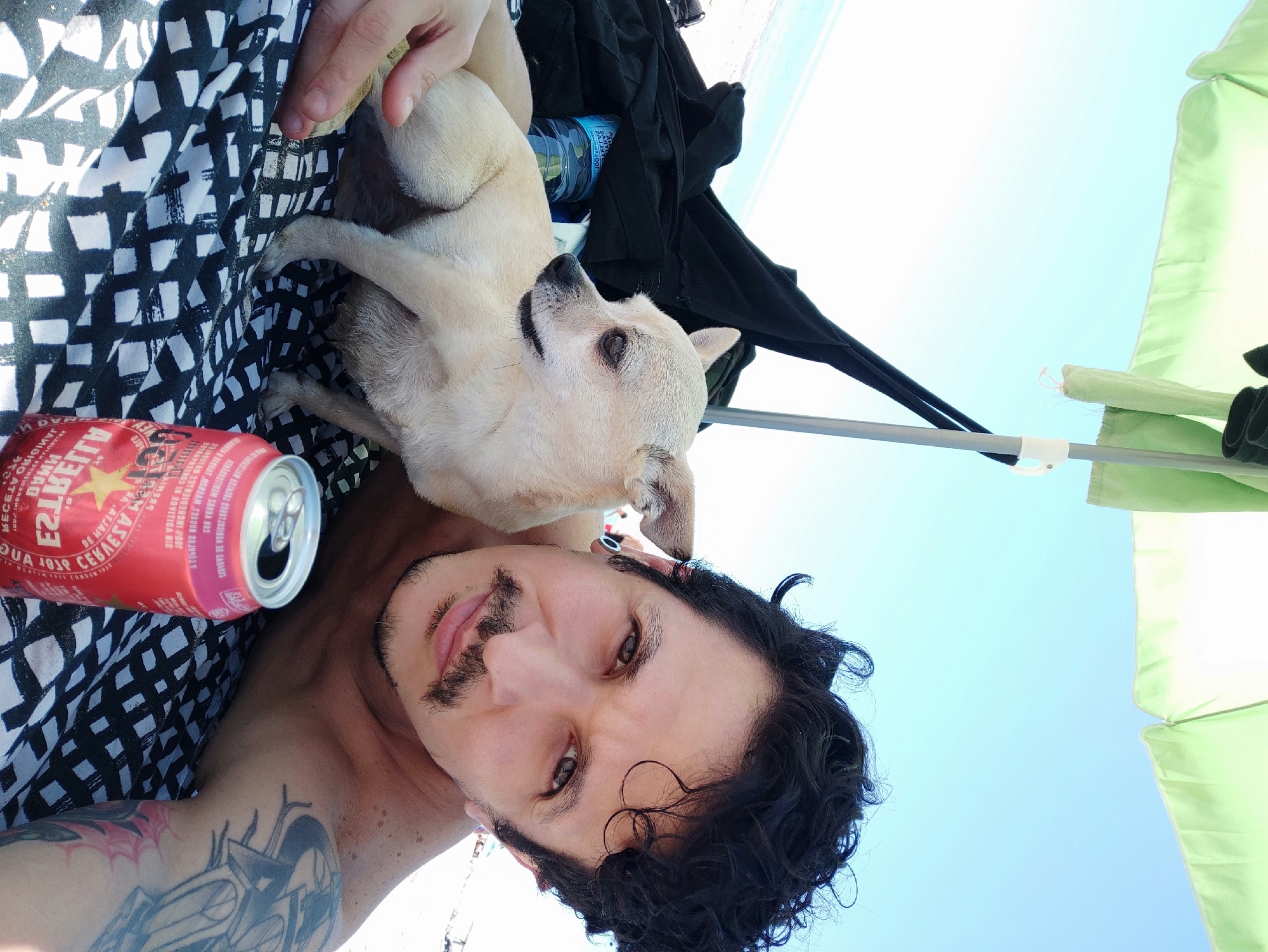 A photograph captures a selfie of a man with his dog outdoors, presumably in Castelldefels, Spain. The man, who appears to be in his late 20s to mid-30s, has dark hair, a mustache, and light skin. He has a tattoo on his left arm. He is looking at the camera with a slight smile. He is shirtless, and there is a can of beer next to him. The dog, a small, light-colored breed such as a chihuahua, is nestled on his chest. The background consists of a blue sky, a beach umbrella, and clothing. The scene appears to be a sunny day at the beach.