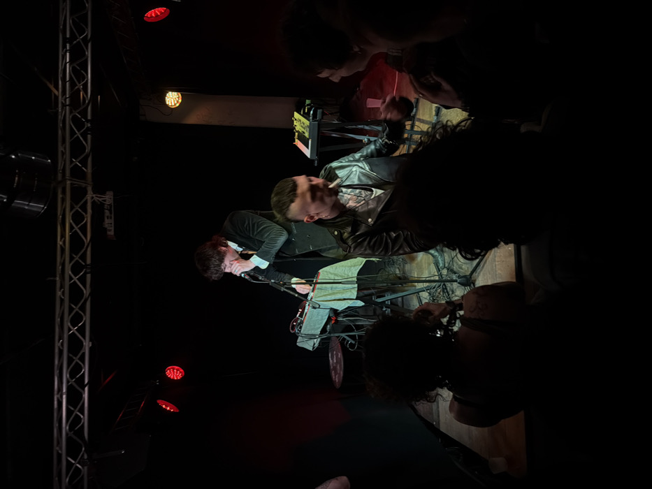 An indoor live music event is depicted within a dark venue in Paris, France. A male performer, with dark, shaggy hair and wearing a dark velvet-like jacket over a light shirt and tie, stands on a raised stage centrally. He holds a microphone to his mouth, appearing to sing or speak. To his left is a stand holding a synthesizer or mixing desk, draped with a light-colored cloth. To his right, another keyboard or synthesizer rests on a stand. The stage is illuminated by overhead truss lighting, including multiple red LED fixtures and a warmer amber light source to the right.
In the mid-ground, slightly off-stage to the right of the performer, a male individual with a mohawk-style haircut and visible tattoos on his neck and chest is seated or standing. He wears a black leather jacket over a light shirt and holds a lit cigarette in his mouth, exhaling smoke. His right hand is raised, gesturing towards another dimly visible individual to his right, indicating an interaction.
The foreground is occupied by several audience members, primarily visible as dark silhouettes. Their heads and shoulders are discernible, suggesting they are observing the performance. One individual in the bottom left has curly hair, and another person in the bottom right holds a clear plastic cup. The flooring of the venue appears to be wooden. The overall atmosphere is dim, with stage lighting providing the primary illumination.