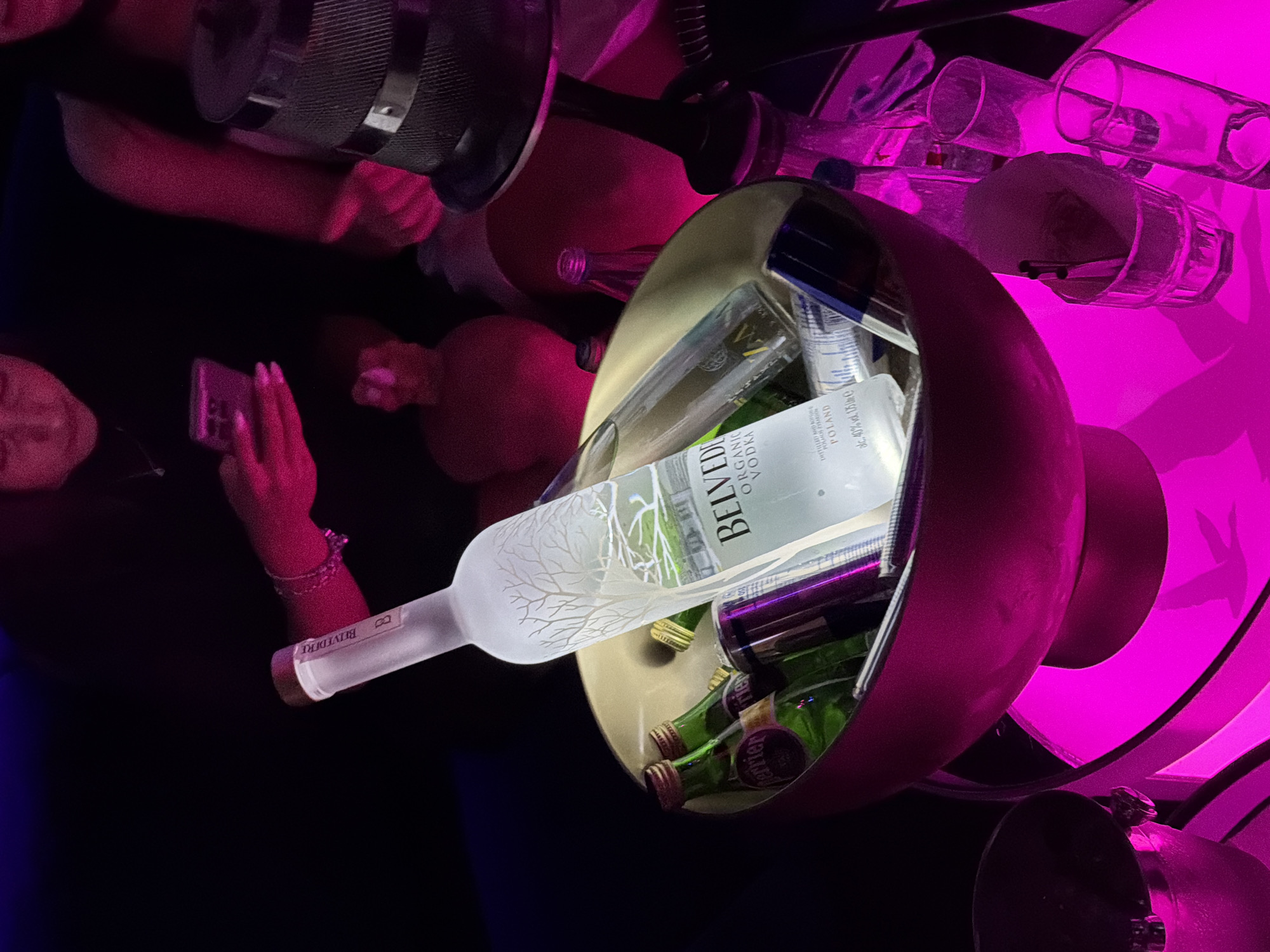 The image shows a bar setting with a Belvedere vodka bottle prominently displayed in a metal container filled with small Perrier bottles and other unidentified items. A person is taking a picture of the display with a smartphone. There are several glasses in the background. The scene is bathed in pink and blue lighting.