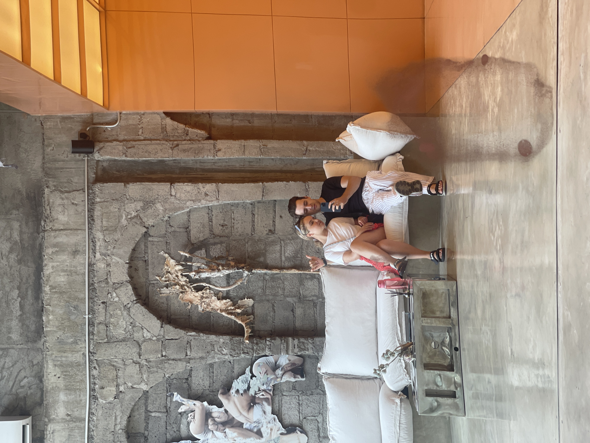 In a lounge area with a grey, rough-hewn stone wall and arched niches, two individuals, a woman and a man, are seated on a white sectional sofa. The woman, on the left, wears a white t-shirt and a red skirt, with her legs crossed. She has blonde hair pulled back and is wearing sunglasses on her head. She is looking down and has her left hand raised in a gesture. The man, on the right, is wearing a black t-shirt and white striped pants. He is holding a smartphone and appears to be showing it to the woman.
In front of the sofa is a metallic, rectangular coffee table with a textured surface and compartments. On the table are decorative branches, a pink water bottle, and a diffuser. To the right of the sofa, a large, orange, paneled wall is visible, with a section of the ceiling featuring illuminated panels. The floor is highly reflective, showing a mirror image of the scene, including the two individuals and the coffee table. The overall aesthetic suggests a modern, art-infused interior space, possibly in a hotel or a cafe, located in the area of North Kuta, Indonesia. The background wall has two large, abstract art pieces depicting human figures, attached to the stone. A black wall-mounted light fixture is visible on the right side of the stone wall.