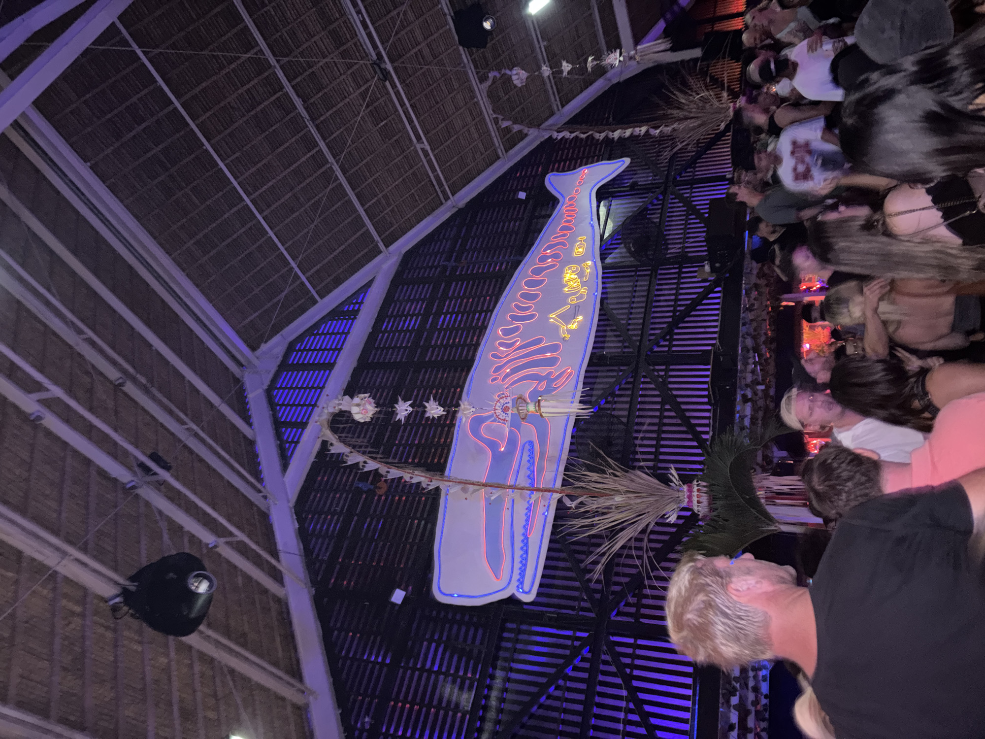 The image depicts a crowded indoor event, likely a bar or club, in North Kuta, Indonesia. A large, illuminated neon sign shaped like a whale dominates the background, set against a dark wall with vertical slats illuminated by purple lights. The ceiling is thatched, supported by white beams and rafters, with spotlights visible. In the foreground, a diverse group of people, predominantly young adults, are gathered, some facing away from the camera and engaged in conversations or looking towards the stage or bar area. The atmosphere appears lively and festive, typical of a nightlife setting.
