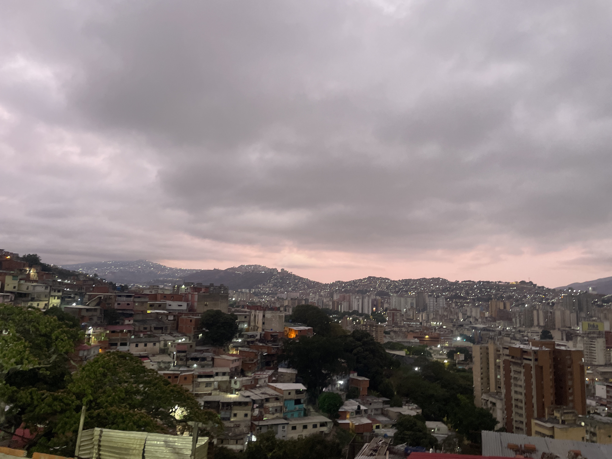 The image depicts a cityscape of Caracas, Venezuela, under a cloudy sky. The foreground shows densely packed buildings and residential areas, typical of the city's informal settlements. In the middle ground, there's a mix of low-rise houses and taller buildings, with patches of green suggesting parks or vegetation. The background features hills covered with buildings, stretching into the distance. The overall lighting is dim, suggesting either dawn, dusk, or an overcast day.