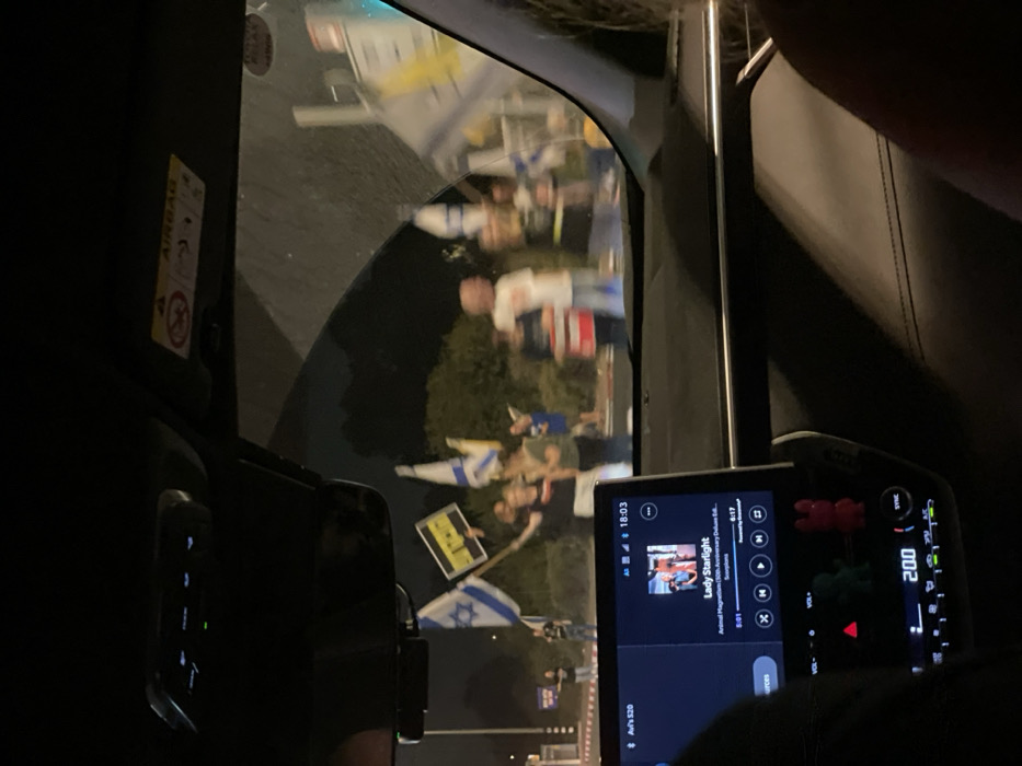 This image captures a nighttime scene from the interior of a vehicle, looking through the windshield at a gathering of individuals on a roadside in Mishmar Hasharon, Israel.
Inside the vehicle, the dark dashboard features an illuminated infotainment screen displaying "Lady Starlight" by "Scorpions," with the time "18:03" visible. Below the screen, the climate control reads "20.0," and two small decorative figures, one green and one red, are positioned. An overhead console is visible, along with a passenger-side sun visor bearing an "AIRBAG" warning label. A person's head and hair are partially visible in the lower right foreground.
Outside the vehicle, approximately 10 to 15 individuals are congregated on what appears to be a paved area or median strip beside a roadway. Multiple individuals are holding flags, predominantly Israeli national flags characterized by a blue Star of David and blue stripes on a white background. One person in the central-left holds a yellow flag with black Hebrew text. The individuals are casually dressed. Some are depicted with raised arms. The background consists of dark foliage. Partial views of street signs or traffic infrastructure are present on the right side of the frame.