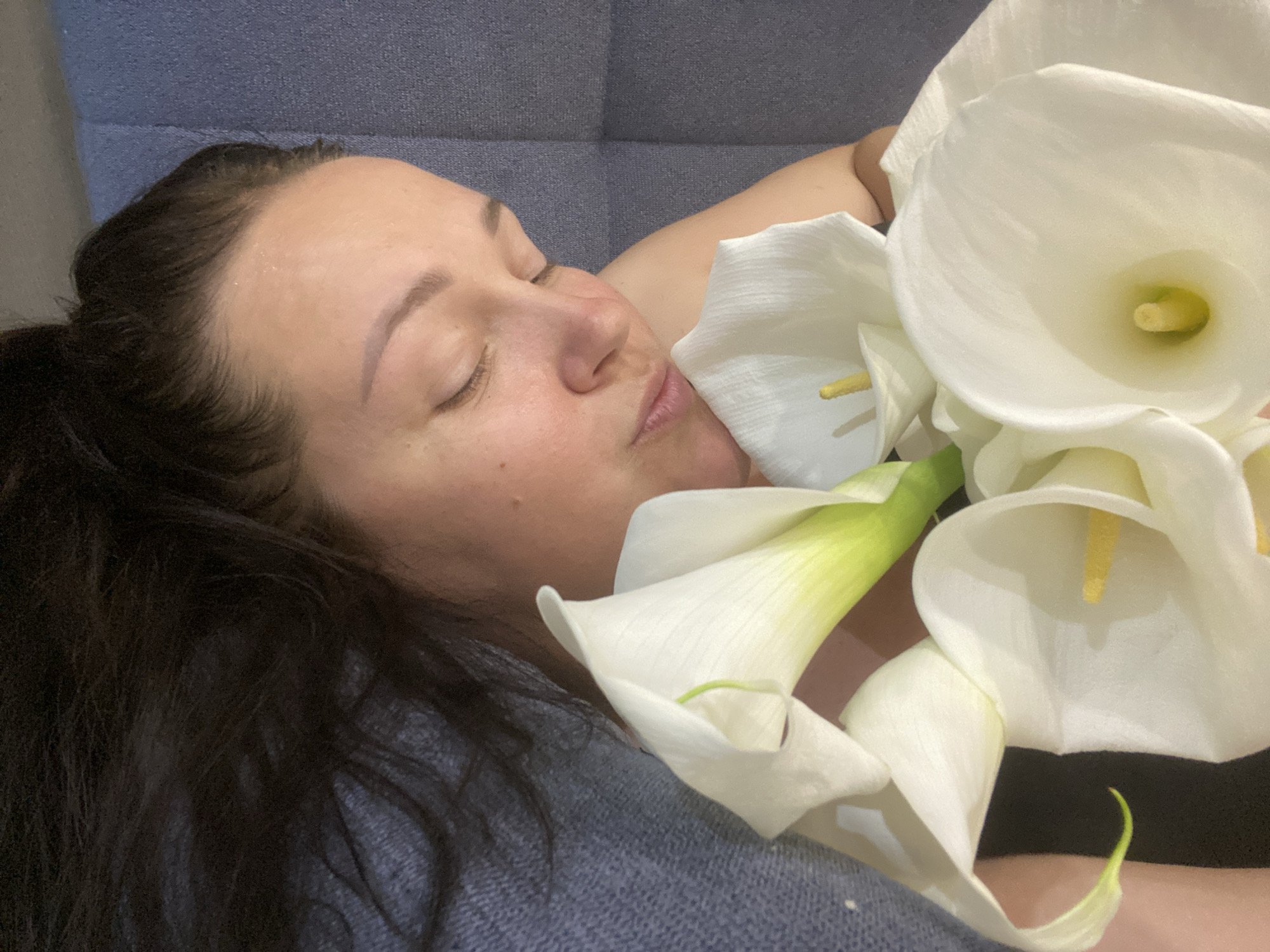 A close-up, high-angle image depicts a female adult with dark hair and light skin, resting her head on a textured grey-blue surface, likely upholstery. Her eyes are closed, and her face is positioned close to a bouquet of white calla lilies. Her right arm/shoulder is partially visible beneath the flowers. Several calla lily blooms are prominent, each featuring a white, funnel-shaped spathe and a visible yellow spadix. Green stems are also discernible. A dark, textured fabric, consistent with clothing, is visible on the lower left section of the frame. The specific city of Sochi, Russia, cannot be verified from the visual content of the image.