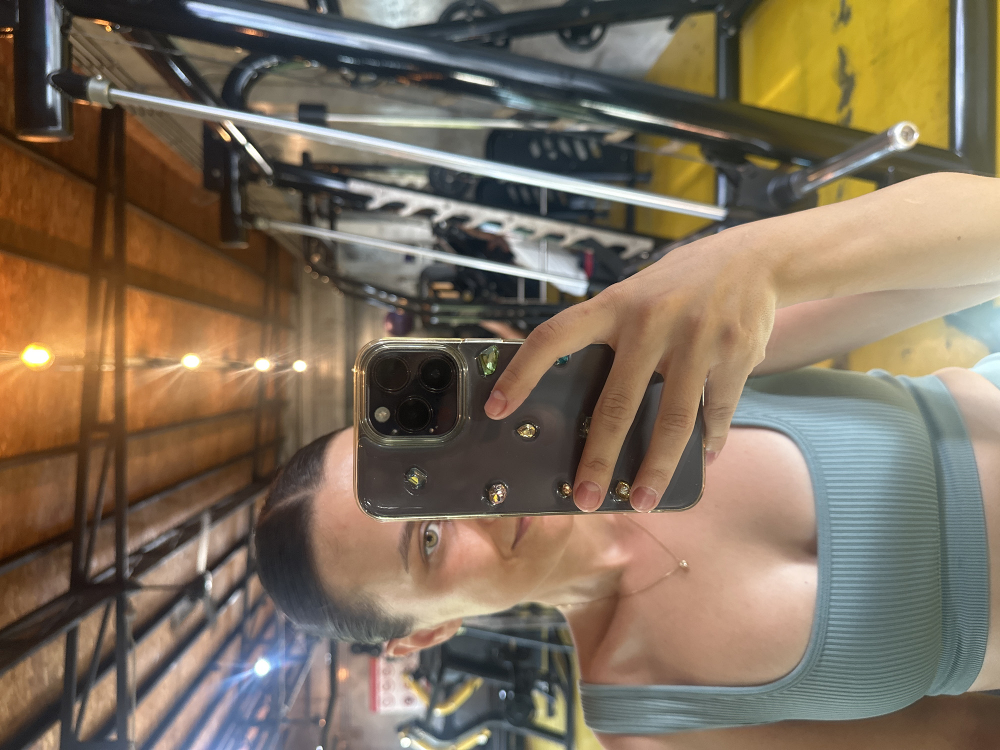 The image is a slightly blurry, vertically oriented selfie taken in a gym. A woman is holding a smartphone with a decorative case in front of her face, capturing her reflection. She is wearing a light blue ribbed tank top and a delicate necklace. Her hair is pulled back, and she has a focused expression.
The background of the reflection shows gym equipment, including metal bars and what appears to be weight machines. The lighting in the gym is warm, with string lights visible, suggesting it might be evening or dimly lit. The overall impression is a casual, personal moment captured during a workout. The location context "Baan Mae Nam, Thailand" provides a geographical setting.