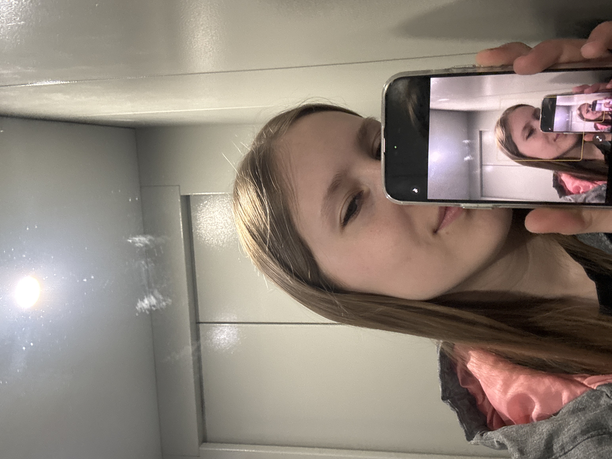 A young, light-skinned person with long, light brown hair is holding a smartphone in their right hand, taking a picture of themselves in a small, white elevator. The elevator has visible smudges and imperfections on its walls, illuminated by a ceiling light. The phone's screen shows a recursive image of the person taking a photo with another phone. The person is wearing a grey jacket with a pink lining. The location appears to be a standard elevator cabin, and without further information, its specific location in Chaikovsky, Russia, cannot be determined.