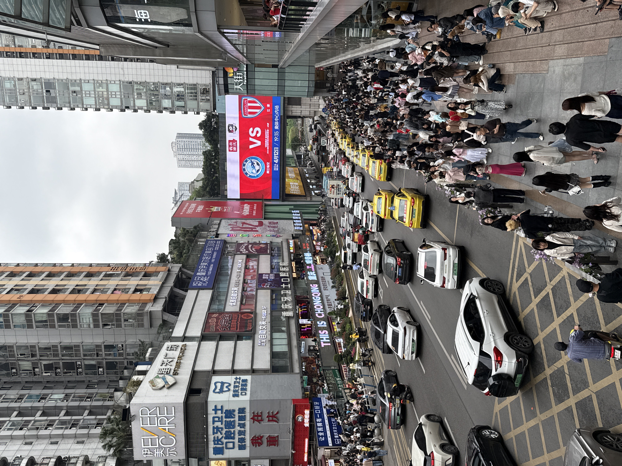 The image captures a bustling street scene in Liangjiang, China. A large crowd of people are gathered on the right sidewalk, partially spilling into the street. The street is filled with various vehicles, including yellow taxis and white cars. Billboards featuring advertisements and notices line the buildings on the left side of the street. A particularly large screen shows a match between Jiangjin and Chongqing. The architecture is a mix of high-rise buildings. Pedestrians are scattered throughout the street. The sky is overcast, indicating potentially gloomy weather conditions.