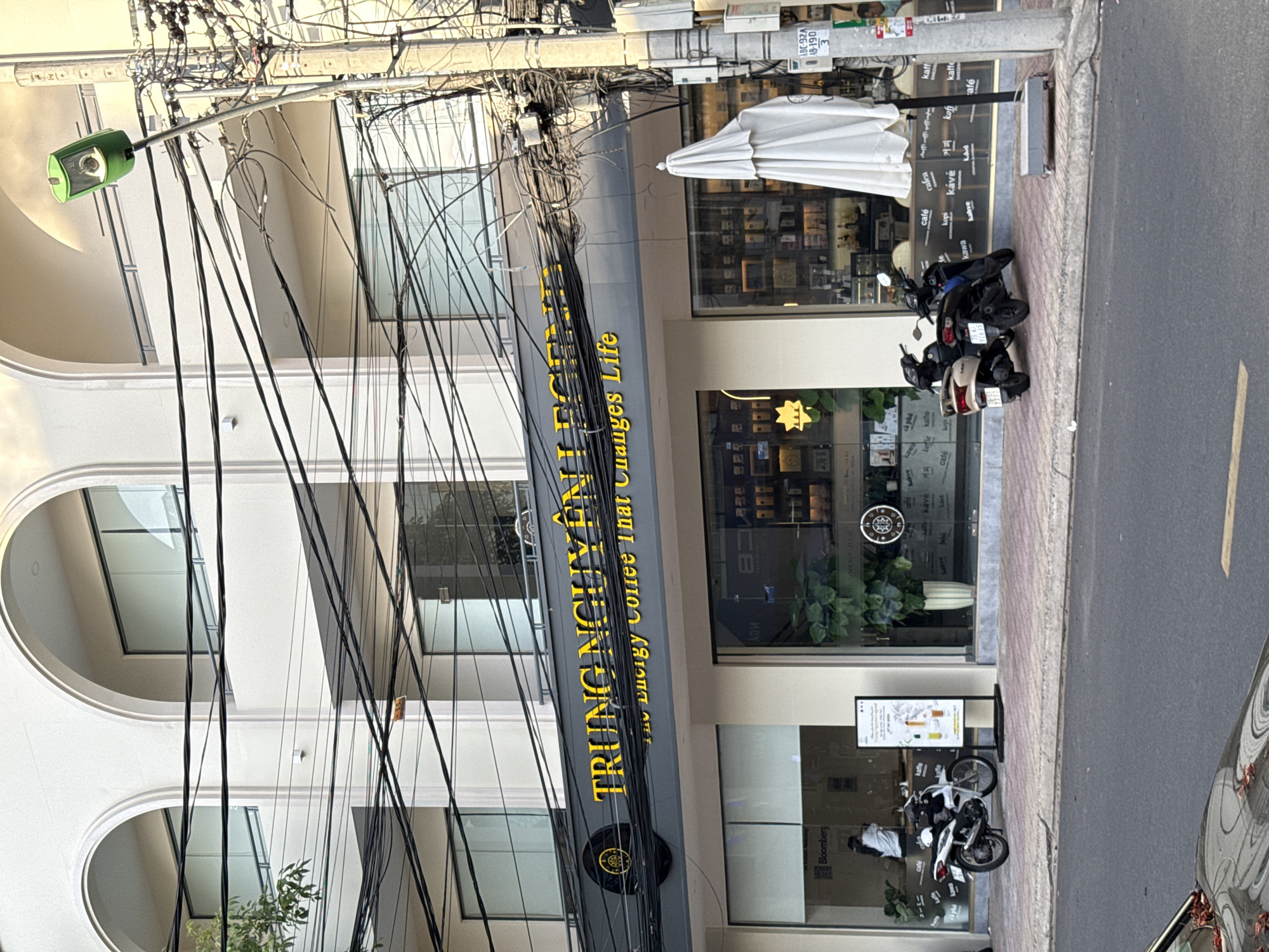 The image captures a street scene in Nha Trang, Vietnam, focusing on the exterior of a Trung Nguyen Legend coffee shop. The building features a modern facade with large arched windows on the upper levels. Below, the ground floor is characterized by a dark-colored awning with the coffee shop's name prominently displayed in yellow lettering. The awning also has a circular logo on the left side.
The storefront windows reveal the interior of the coffee shop, showcasing displays of coffee products and indoor plants. Several motorcycles are parked along the sidewalk in front of the shop. A tall, slim white umbrella is positioned near the right side of the entrance, partially obscuring the view of the window. A street lamp with a green housing is visible on the right side of the image, attached to a utility pole laden with numerous electrical wires. The sidewalk is paved and appears to be well-maintained. The street in front of the sidewalk shows a portion of a car and lane markings. The overall environment suggests a typical urban setting in Vietnam.