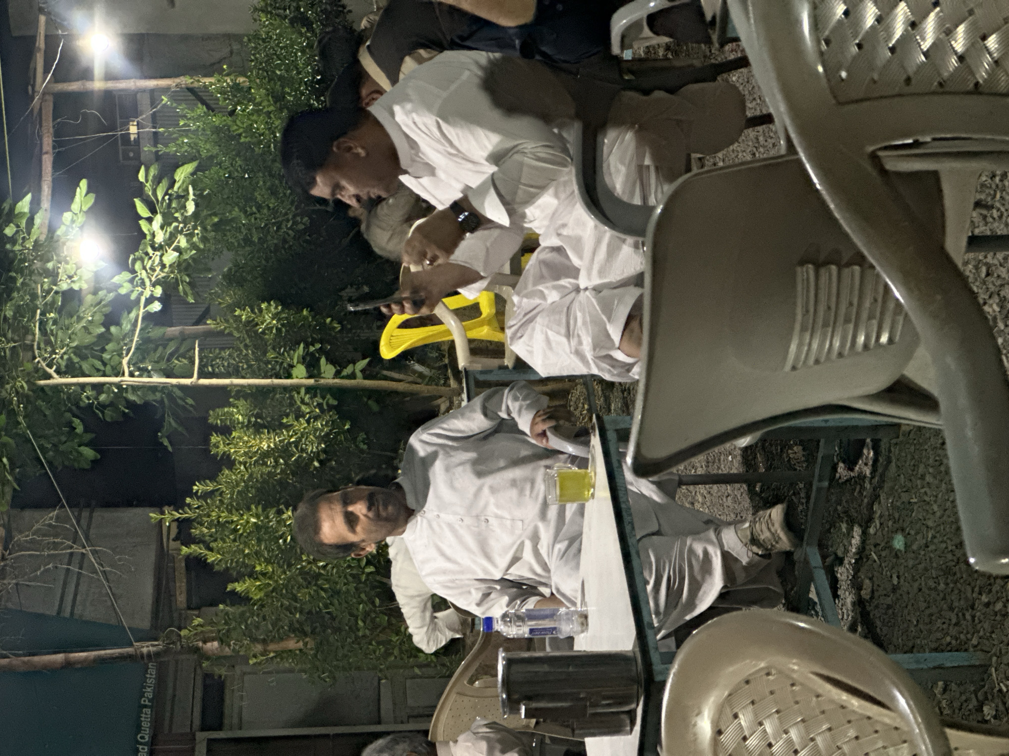 The image depicts an outdoor night scene featuring a casual gathering of men. Two men, prominently in the foreground and mid-ground, are seated at tables wearing traditional white shalwar kameez. The man in the foreground, with a mustache, looks towards the viewer while seated at a light-colored table with a glass of yellow beverage and a plastic water bottle. Behind him, another man with a smartwatch on his left wrist is focused on his mobile phone. Partial views of other individuals and chairs suggest a larger gathering.
The setting is an informal outdoor area, possibly a garden restaurant or courtyard, illuminated by several bright artificial lights. Lush green bushes and trees form a dense background, reflecting some of the light. The ground is covered with gravel or small stones. Various simple plastic chairs, some with woven patterns on their backrests, are arranged around the tables.
The time of day is clearly night, as indicated by the artificial lighting. A faint watermark or text "Quetta Pakistan" is visible in the bottom left corner, providing a location context for the scene. The overall ambiance appears relaxed and social.