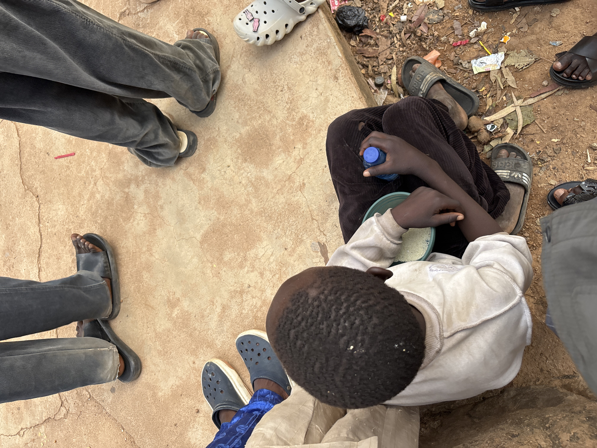 In Jos, Nigeria, a close-up shot captures the lower bodies and immediate surroundings of several individuals. A young boy, seated on the ground, is the central focus, wearing a light-colored sweatshirt and dark brown trousers. He is holding a blue water bottle in his right hand and has a bowl of what appears to be food in front of him. His feet are bare. Around him, other individuals are visible, indicated by their legs and footwear. One person is wearing dark blue Crocs, while another has on dark grey, baggy corduroy trousers and black sandals. Further to the right, a pair of grey Crocs with pink accents are visible on the ground. The ground surface is a mix of concrete and dirt, with scattered leaves and debris. The overall impression is a snapshot of everyday life in a communal setting.