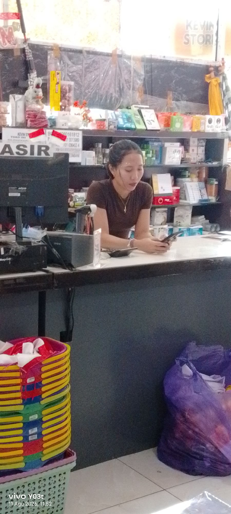 A young woman with dark hair pulled back and wearing a brown t-shirt is seated behind a counter in a store. She is looking down at a mobile phone in her hands. The store appears to be a retail establishment, possibly a convenience store or small grocery store, with shelves stocked with various goods in the background. There is a computer monitor on the counter to her left, and a basket filled with colorful items, possibly plastic containers, to her left in the foreground. A large purple plastic bag filled with items is on the floor to her right. The lighting suggests an indoor setting, possibly during daytime. The image also contains a timestamp and device information at the bottom left corner: "vivo Y03t 11 Agu 2026, 11.52".