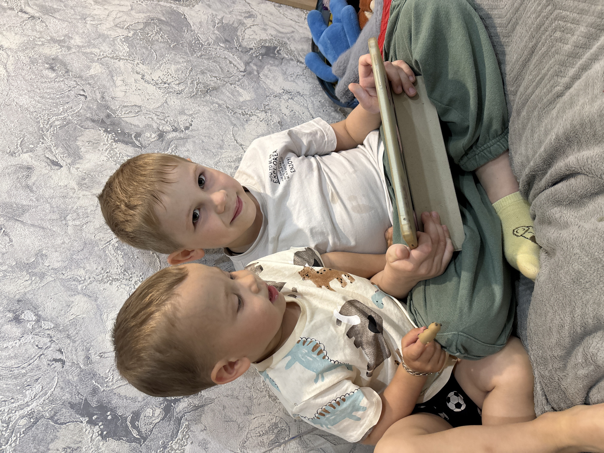 This media file captures two young boys relaxing indoors in Rusanovo, Russia, during what appears to be daytime. The children are lying down on a light grey, textured surface, possibly a blanket or rug, with an additional plain grey blanket visible in the background.
The older boy, with light brown hair, is seen on his side, wearing a white short-sleeved t-shirt, green sweatpants, and light yellow socks. He holds a tablet or large smartphone, encased in a light beige cover, with both hands. He is looking directly at the camera with a warm, smiling expression. His t-shirt bears the visible text: "HOW TO BE AN EXPLORER FIND SOME AD INVITE YOUR FRIEN BE IN A GOOD MOOD ENJOY!"
Beside him, the younger boy, also with light brown hair, is lying down, dressed in a light-colored t-shirt patterned with blue dinosaurs and brown animals, and black shorts or underwear featuring a white soccer ball design. He holds a small, light-brown object, possibly a biscuit or a wooden toy, in his right hand and has a silver-colored bracelet on his wrist. His gaze is directed downwards or to the side, appearing focused on his activity or the object he is holding.
In the background, a blue plush toy is partially visible, indicating a casual, comfortable indoor setting. The image portrays a relaxed moment shared between the two siblings.