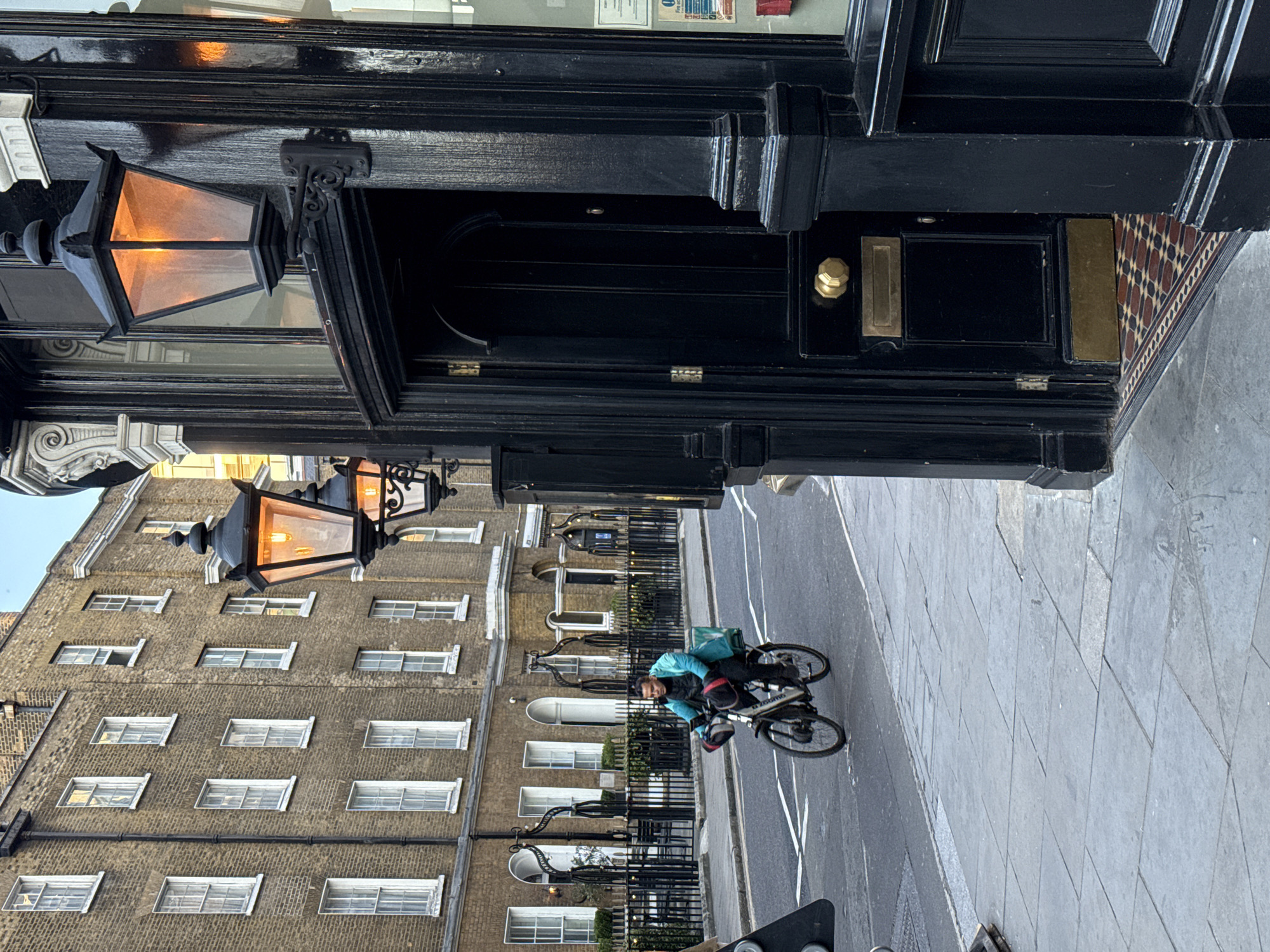 The image depicts a street scene in London, United Kingdom. In the foreground, there is a black doorway with a brass doorknob and mail slot. Above the doorway, a lit gas lamp is mounted on the building's facade. Across the street, a cyclist wearing a light blue jacket rides in the road. The cyclist appears to be making eye contact with the camera. A brick building with multiple windows stands in the background behind a black wrought-iron fence. The road surface is paved with stone.