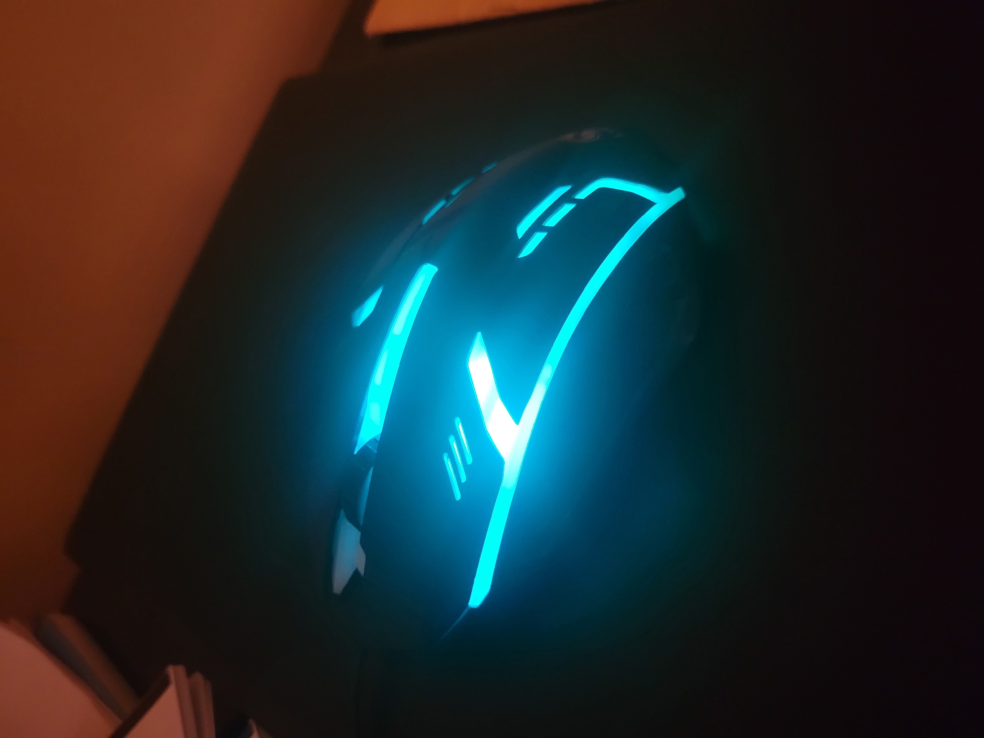 An illuminated computer mouse sits on a dark surface, likely a desk or mousepad. The mouse is predominantly black with glowing cyan accents in various geometric patterns and lines, including three parallel lines on its surface and a larger, angular design along its side and front. The lighting from the mouse casts a bright cyan glow on the surrounding dark area. The background is out of focus and appears to be a dark surface with a hint of warm, orange light from above, possibly from a lamp or ambient room lighting. There are no people or animals visible in the image, and no specific location details are discernible.