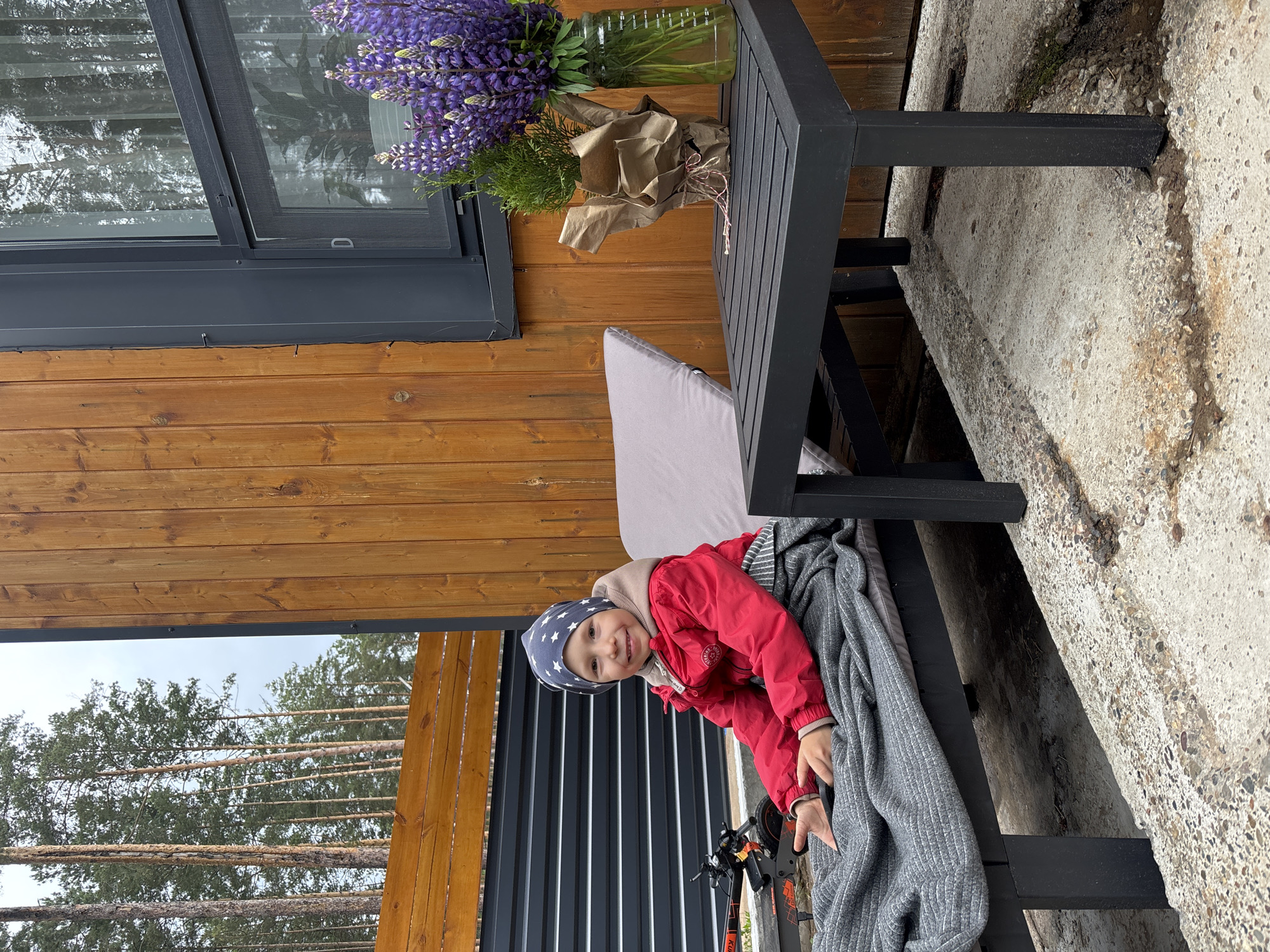 This image captures an outdoor patio scene in Chaykovsky, Russia. A young child, smiling and looking directly at the camera, is seated on a grey outdoor lounge chair, covered from the waist down by a ribbed grey blanket. The child wears a red jacket, a light brown hooded sweatshirt, and a dark grey beanie adorned with white star patterns. To the child's left, partially visible, is a dark electric scooter or bike with orange accents.
To the child's right, a dark grey slatted outdoor table holds two items: a clear glass vase containing a bouquet of vibrant purple lupine flowers, with measurement markings visible on its side, and a small potted evergreen plant wrapped in brown kraft paper, tied with red and white string.
The background features a building with vertical wooden paneling and a dark grey window frame, which reflects tall pine trees. Behind a dark grey slatted fence and a wooden barrier, a forest of slender pine trees extends upwards towards an overcast sky. The ground surface is rough concrete, showing some minor discoloration and unevenness.