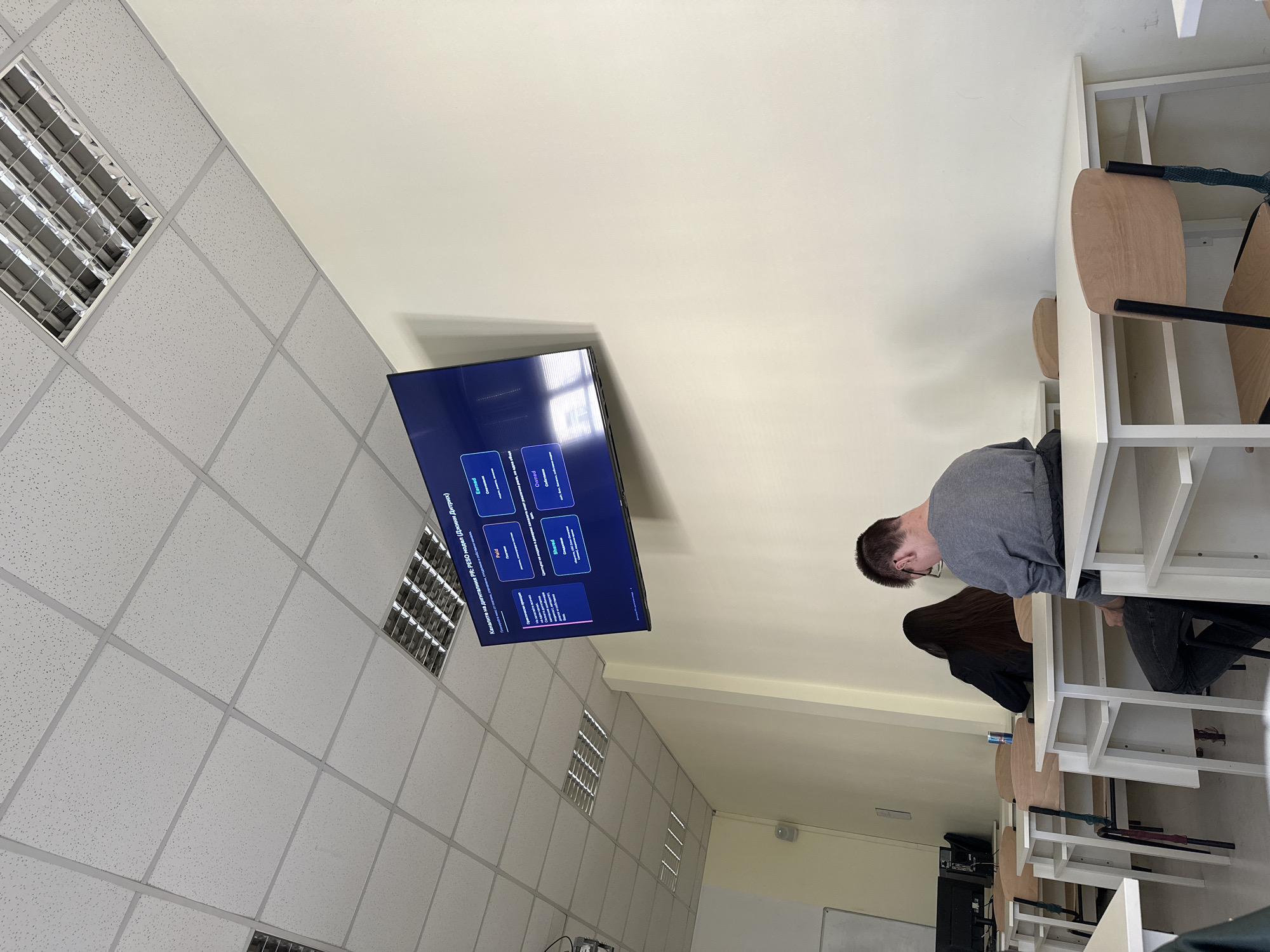 The image depicts a classroom setting in Sofia, Bulgaria. A flat-screen display is mounted on the wall, showing a presentation slide, possibly related to software or technology. Two individuals are seated at desks, attending to the material. One is wearing a grey long sleeve shirt. The classroom is lit by recessed fluorescent lights in a drop ceiling. Desks and chairs are neatly arranged in rows. The color palette is neutral with a yellow tone, giving a well-lit atmosphere. The setting suggests an educational or training environment.