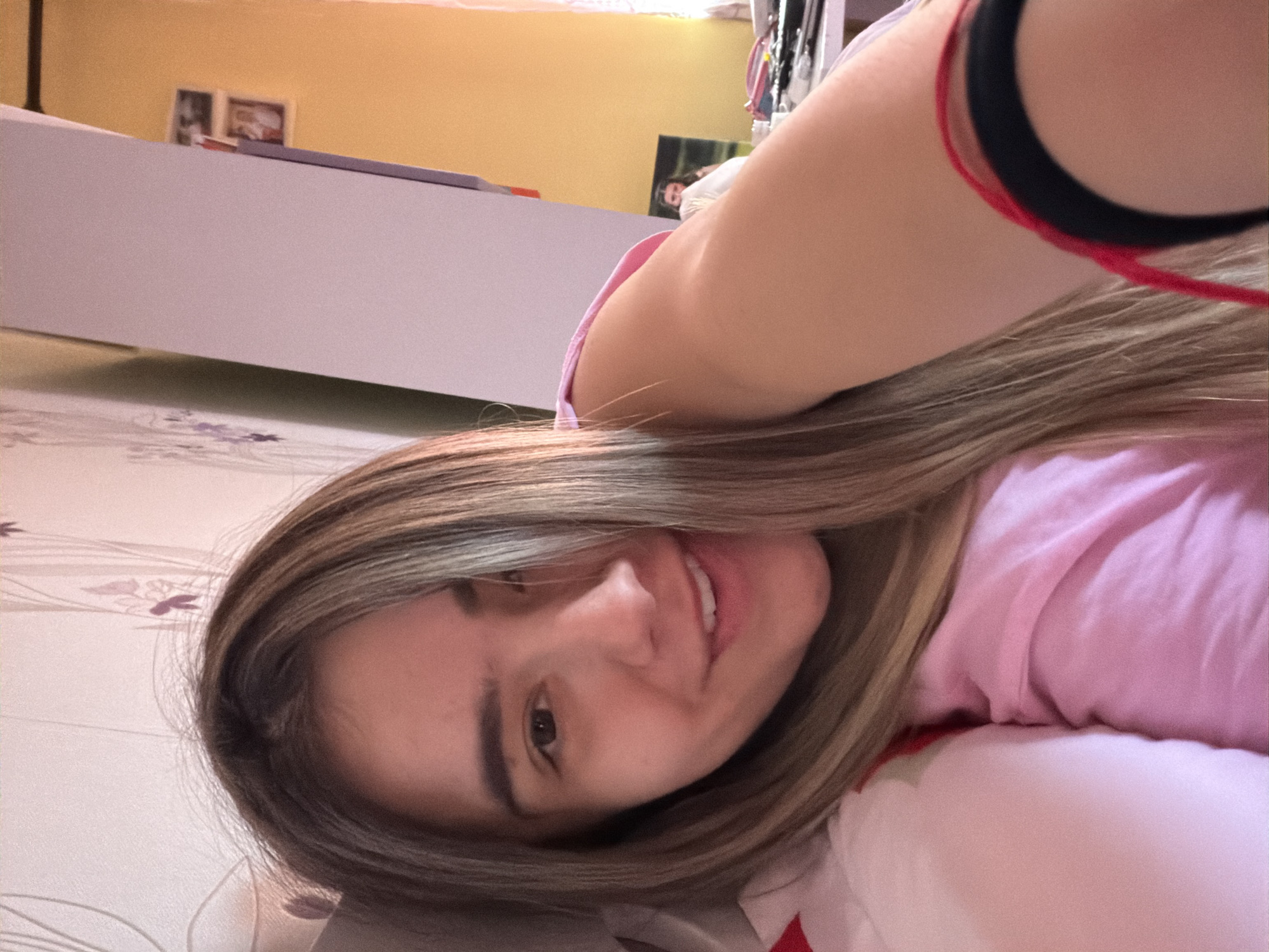 A woman is lying down, likely on a bed or similar soft surface, indoors. She is wearing a pink top. She has light brown hair with blonde highlights. She is smiling slightly. A black and red wristband is visible on her arm. The background includes a white piece of furniture, a yellow wall, and various objects that appear to be in a bedroom. The image is taken from a close-up angle, focusing on her face and upper body. Location is not identifiable.