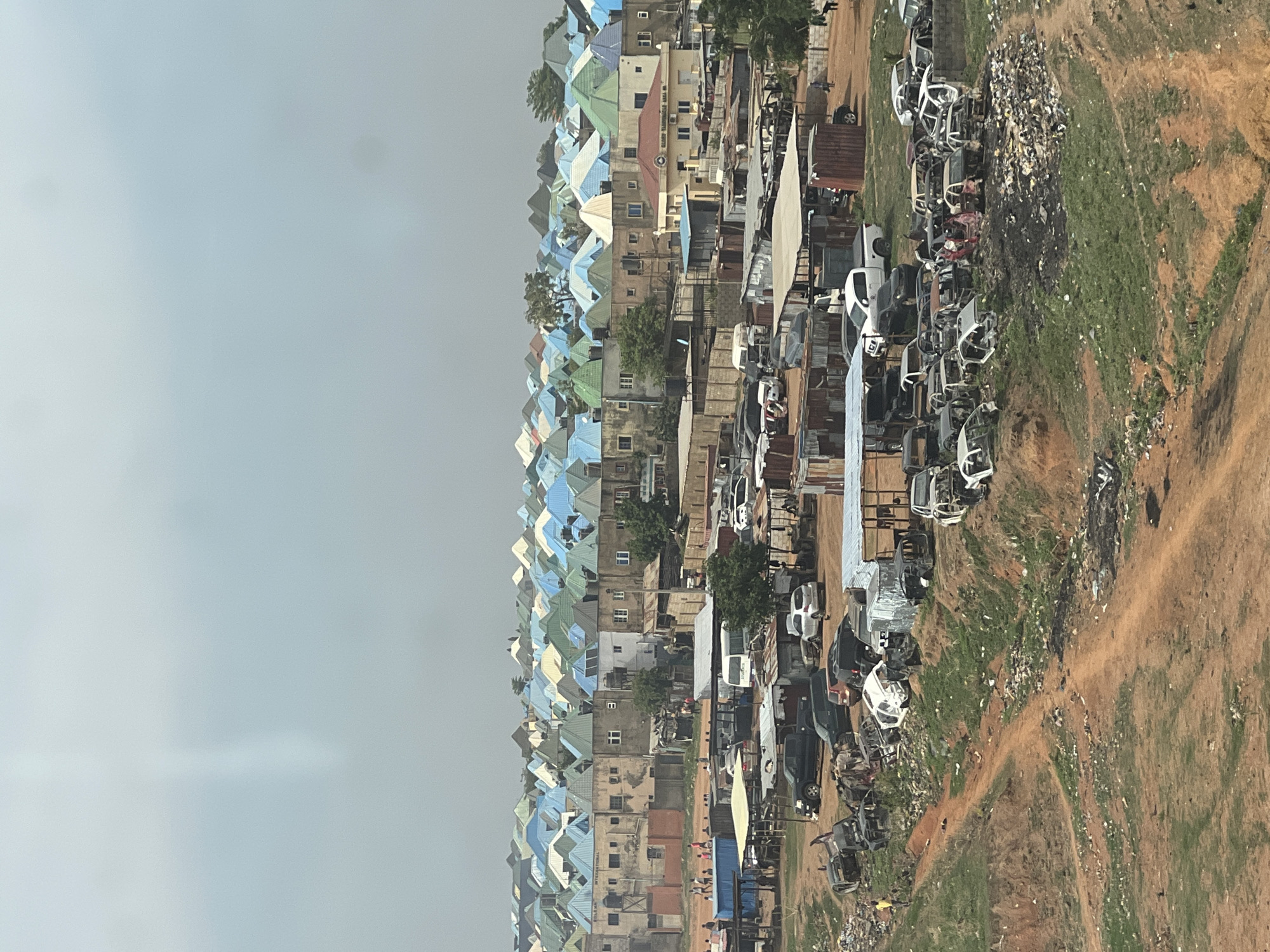 The image shows an aerial view of a section of Abuja, Nigeria, characterized by a mix of residential and commercial activity. On the left, closely packed houses with predominantly blue, green, and light-colored roofs dominate the landscape. Adjacent to the houses, there appears to be a commercial area or junkyard with numerous vehicles and vehicular parts scattered across the earth. Further to the right, the vegetation is sparse and the ground is a mix of dirt and grass. The sky is overcast.
