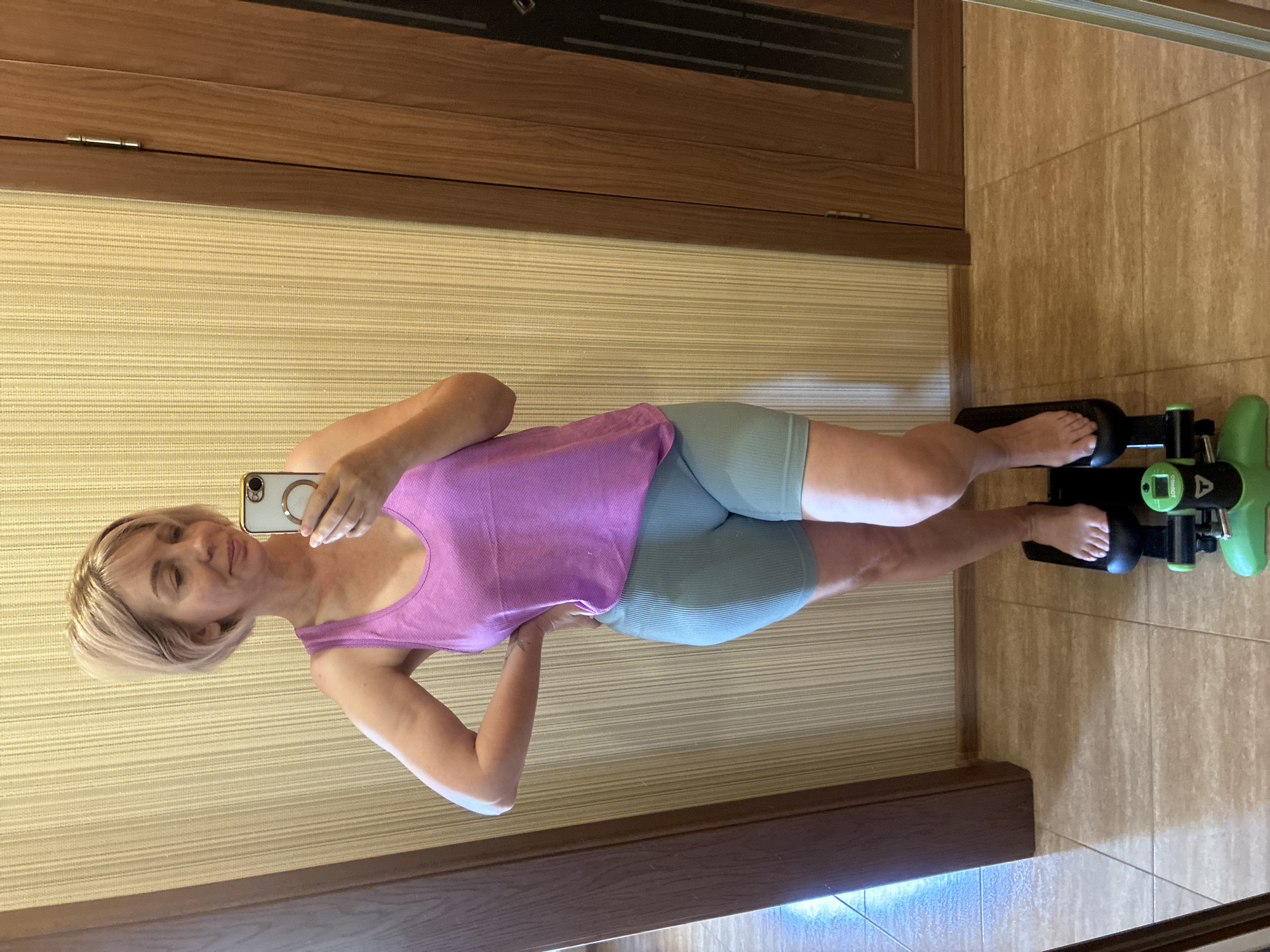 The image captures a fair-skinned woman, approximately middle-aged, taking a mirror selfie indoors. She has short, light blonde hair styled in a bob and is looking towards her reflection with a slight smile. She is wearing a light purple tank top and light teal, ribbed biker shorts. Her right hand rests on her hip, while her left hand holds a white smartphone with a gold ring holder on the back, angled to capture the reflection. She stands barefoot on a compact black and green exercise machine, identified by the visible "OMNEO" logo on its green console. The background features a light beige wall with a subtle vertical striped texture. On the right, a wooden door with a dark paneled window is visible, and on the left, the dark brown edge of another doorway or furniture piece is present. The floor is covered with light brown tiled flooring. No visual cues within the image definitively confirm the location as the city of Sochi, Russia.