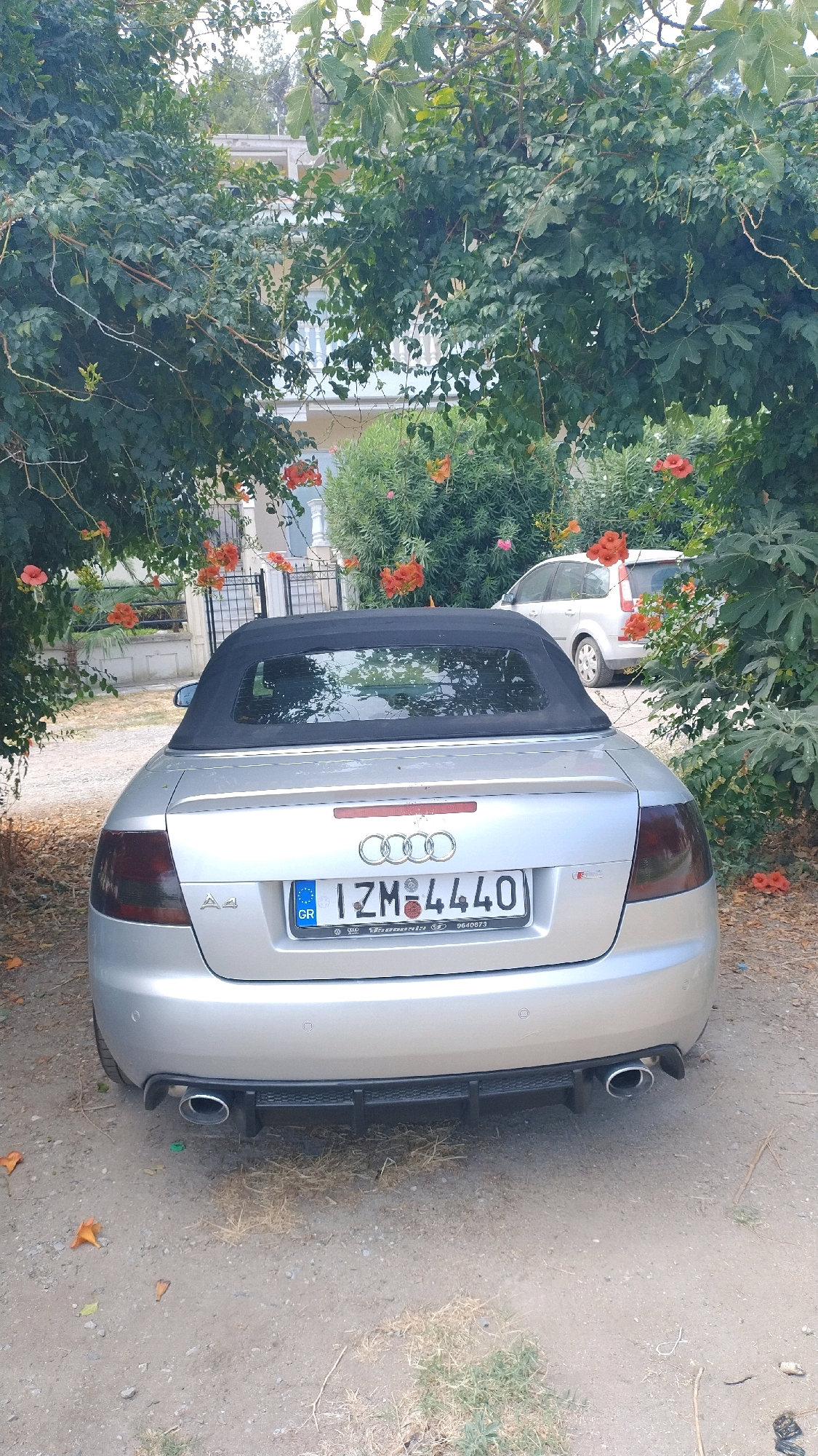 An overturned silver Audi A4 convertible is parked on a dirt path in front of a house in Agia Triada, Greece. The car is facing away from the camera, and its black convertible top is up. The car's license plate is visible and reads "IZM 4440". The car is surrounded by trees and bushes, some of which are bearing orange flowers. In the background, a white minivan is parked in a driveway. The house appears to be two stories tall and has a white balcony. The scene is lit by natural daylight.