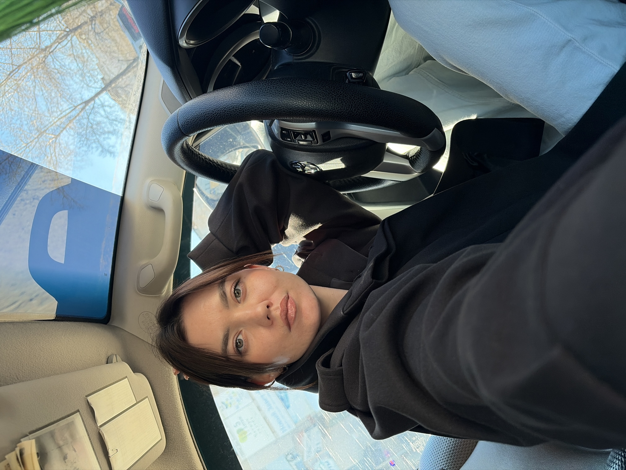 A woman is taking a selfie inside a car. She is wearing a dark hoodie. The steering wheel of the car is visible in the background, along with a street scene through the windshield. The text on a sign outside the car mentions Chaikovsky, Russia.
