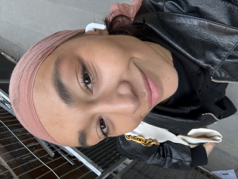 The scene captures a young adult female in an urban outdoor setting. She possesses a medium skin tone, dark eyes, and dark, pronounced eyebrows, with a subtle, closed-mouth smile directed upwards at the viewer. Her head is covered by a light pink or peach-colored fabric headscarf, from which some dark, curly hair emerges at the back. She is attired in a black faux leather motorcycle jacket, featuring a zippered front and a notched lapel. A black garment is visible beneath the jacket. In her right ear, a white wireless earbud is present. A white bag with a large, gold-toned chain strap is visible on her left side. The background consists of indistinct metallic structural elements, possibly scaffolding or temporary barriers, on the upper left, and a plain, light-gray textured surface, likely a wall or ground. The precise location within Paris, France, is not identifiable from the visible environmental details.