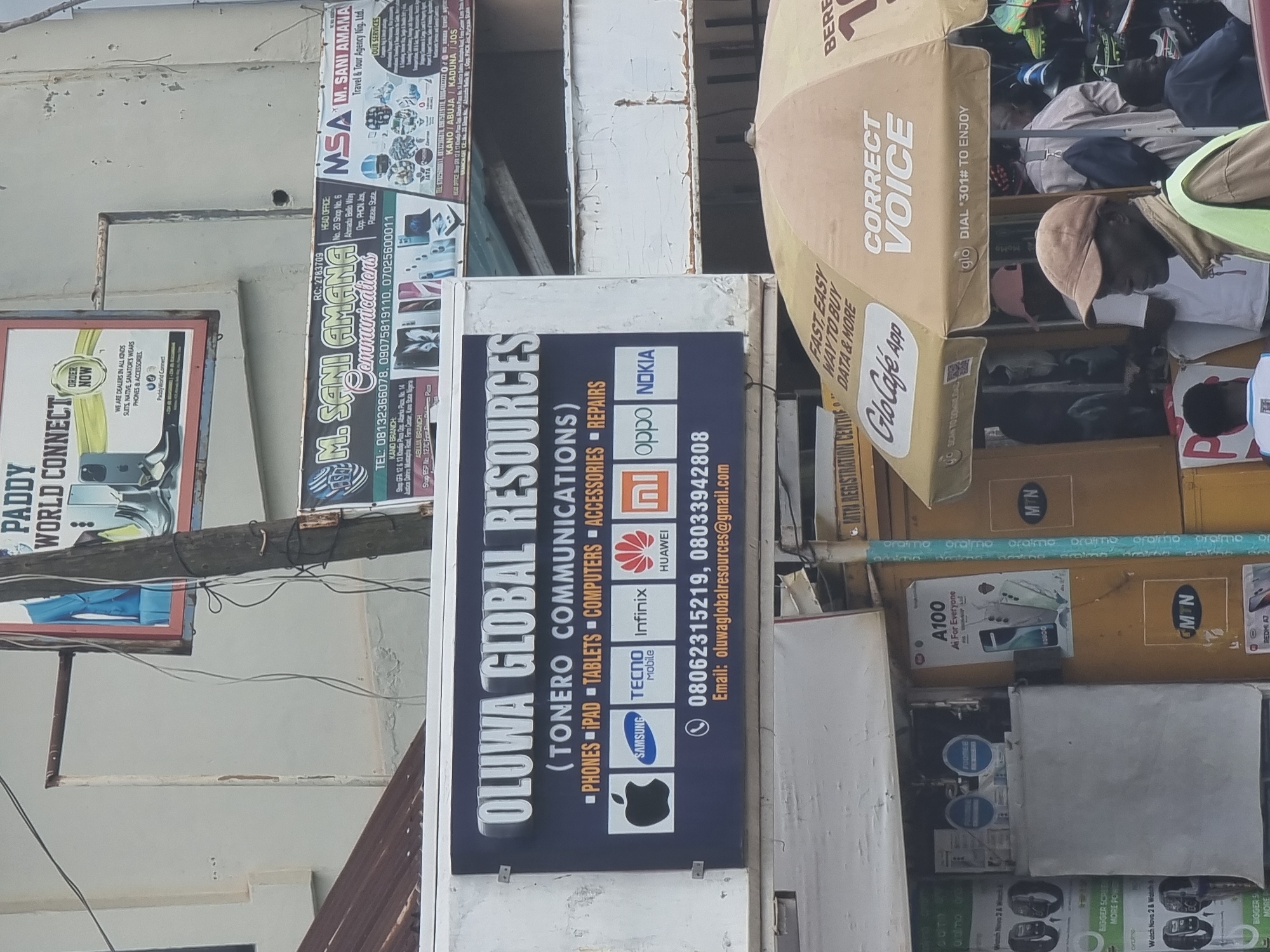 This outdoor shot captures a storefront in Jos, Nigeria. Prominently displayed is a rectangular sign for "OLUWA GLOBAL RESOURCES (TONERO COMMUNICATIONS)," listing phones, tablets, computers, accessories, and repairs, and showing brand logos (Apple, Samsung, Tecno, Infinix, Huawei, Oppo, Nokia) along with contact numbers and an email address. Adjacent to it is a sign for "M. SANI AMANA Communications." The photo is taken in daylight and features an umbrella bearing advertising for Glo Café App and "Correct Voice." Merchandise is displayed in boxes, some branded with MTN, and a person wearing a cap and green reflective vest is partly visible. Other signs are present in the background, including one that says "PADDY WORLD CONNECT." A tangle of wires runs overhead.