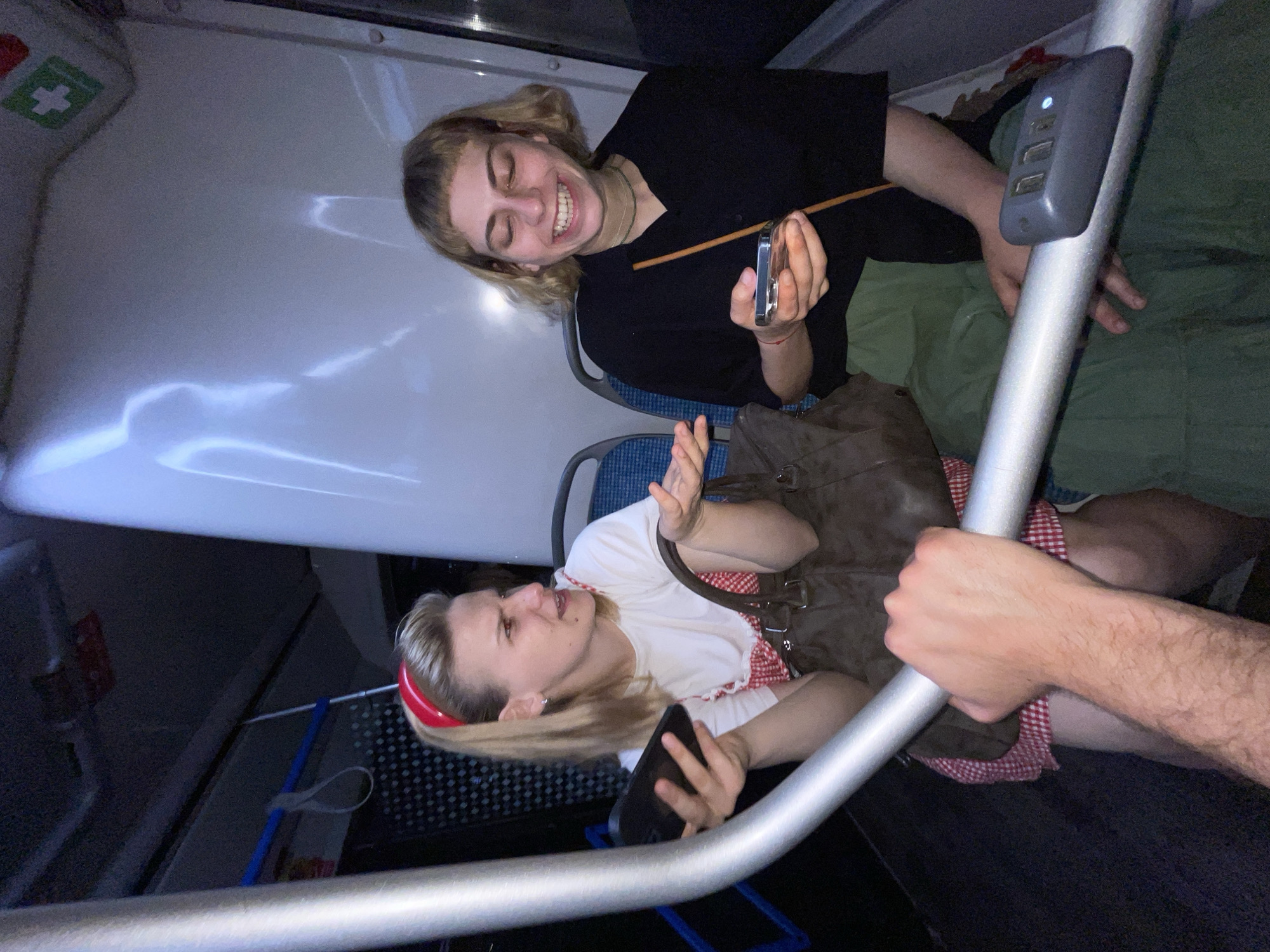Two young women are riding public transportation, possibly a bus, in Moscow, Russia. One woman with short blonde hair is wearing a black shirt and green pants, smiling while holding a silver phone or device. The second woman, also blonde, is wearing a white shirt with a red checkered pattern and a red headband, holding a black smartphone. A brown handbag rests beside her. A man's arm is visible gripping a silver handrail with a USB charging station attached. The interior of the bus is visible, including seats, windows, and safety features.
