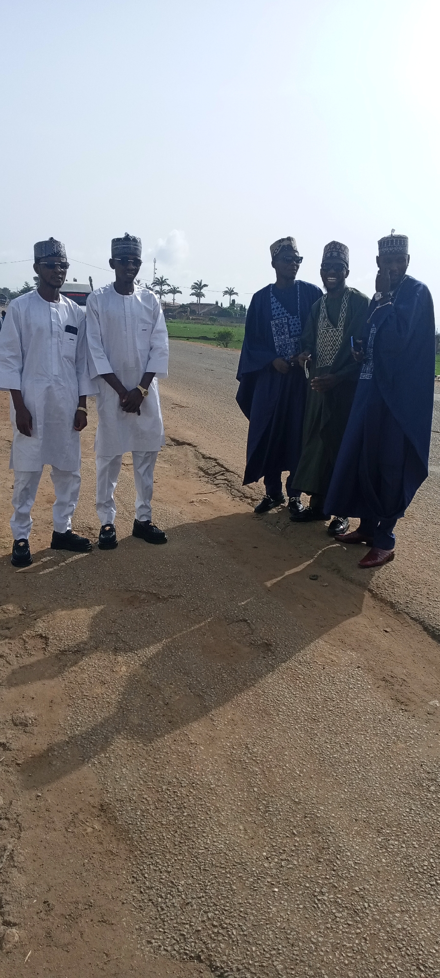 The image depicts five men standing on a paved road. Two men on the left wear matching white outfits and caps, with black shoes. Three men on the right wear dark blue or green outfits and caps, and shoes of varying colors. The background shows vegetation and buildings under a bright sky. The ground is a mix of paved and unpaved surfaces. The location is Naraguta Hausawa, Nigeria.