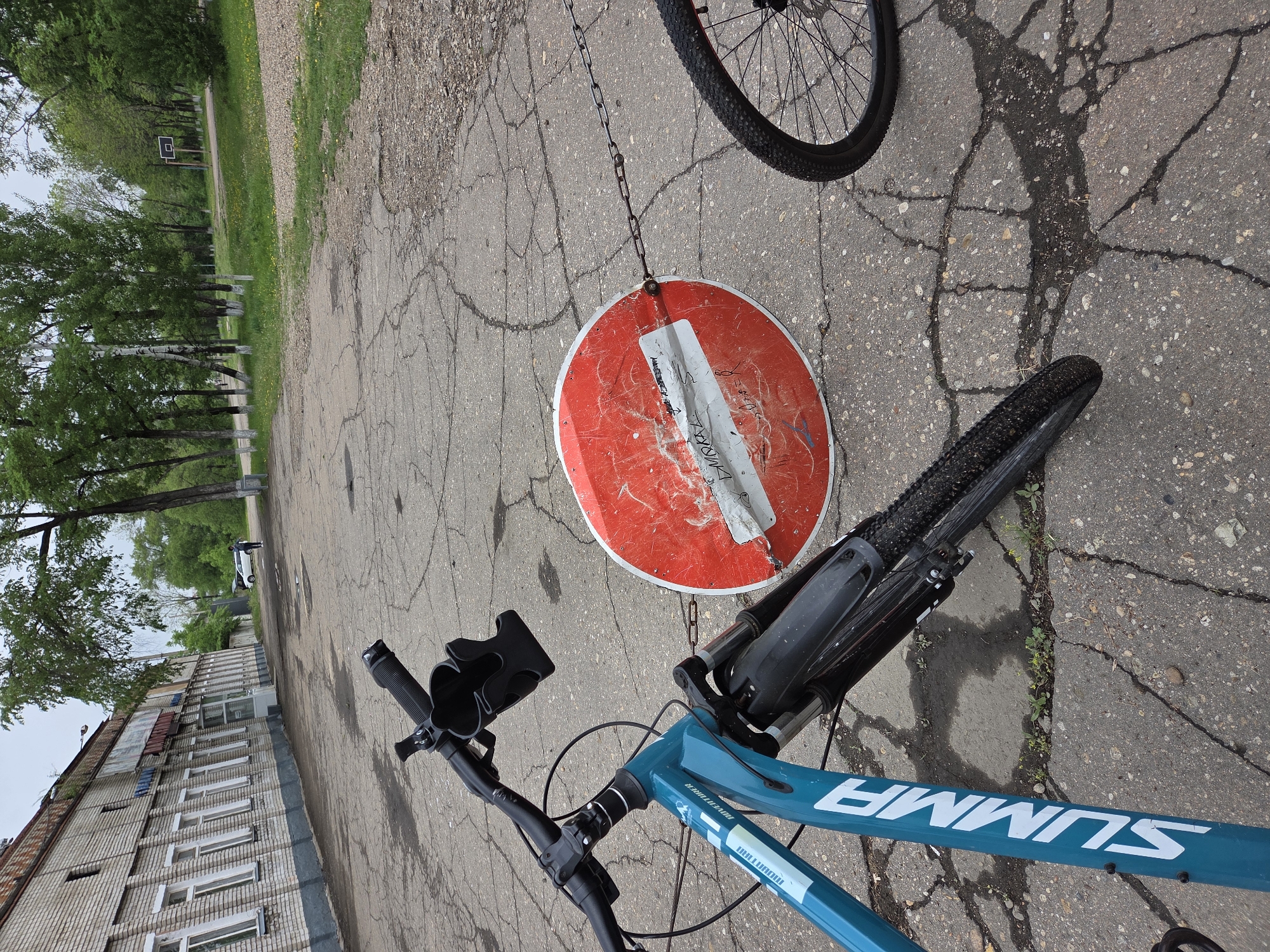 The image depicts a blue bicycle with "SUMMA" written on its frame, positioned on an asphalt surface. A damaged, round, red and white "Do Not Enter" sign hangs by a chain between the bicycle's front tire and handlebars. The asphalt is cracked and shows signs of wear. In the background, there is a multi-story brick building with windows and, further off, trees and a basketball hoop. The location is Birobidzhan, Russia, indicated by the word "SUMMA".