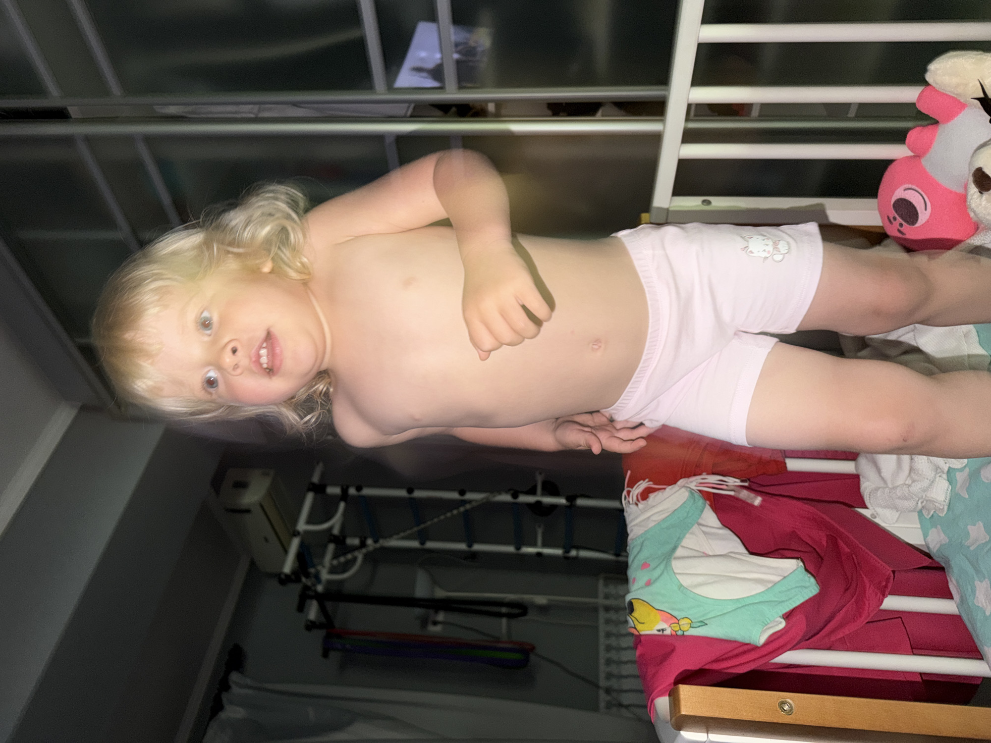 This is a vertically oriented, low-angle, indoor photograph of a young child lying down. The child has blonde hair and is wearing light pink underwear with a small white cat graphic on the left leg. The child's upper body is bare and they are looking upwards towards the camera with a slightly open mouth and a curious expression. Their left arm is bent across their chest.
The child appears to be on a bed with a white railing, possibly a crib or bunk bed. Behind the child, a blurred pink and grey stuffed animal toy is visible, along with other laundry items. A dark, slatted structure is in the background, and a white electrical device is mounted on a wall behind the child's head. The overall lighting is dim, suggesting it might be nighttime or a poorly lit room. The scene appears to be a bedroom or nursery. No visible text is present in the image. The context of Sochi, Russia, is provided, but there are no specific visual cues within the image to confirm this location.