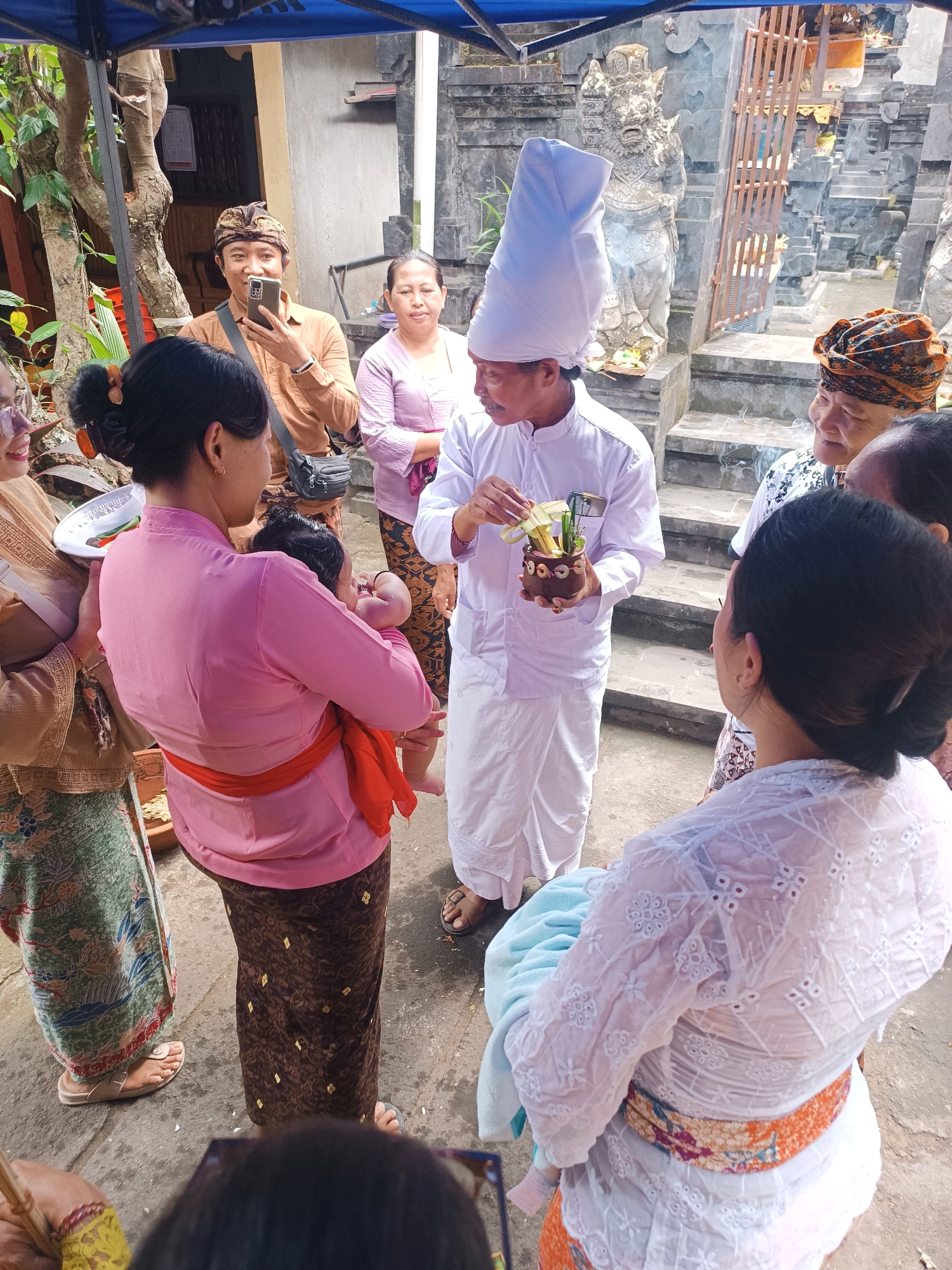 In a courtyard with stone steps and a weathered stone statue of a deity, several people are gathered. A man in a white robe and a tall, white, conical head covering stands center, holding a small brown pot containing greenery and what appear to be offerings. To his left, a woman in a pink blouse and brown patterned skirt holds a baby wrapped in orange fabric. Behind her, a man in a brown shirt holds up a smartphone, seemingly taking a photo. A woman in a purple top stands to the man's right, and another woman in a patterned head covering is visible behind the man in white. The ground is paved with concrete. The scene suggests a religious or cultural ceremony taking place in a temple complex, possibly in Tabanan, Indonesia.