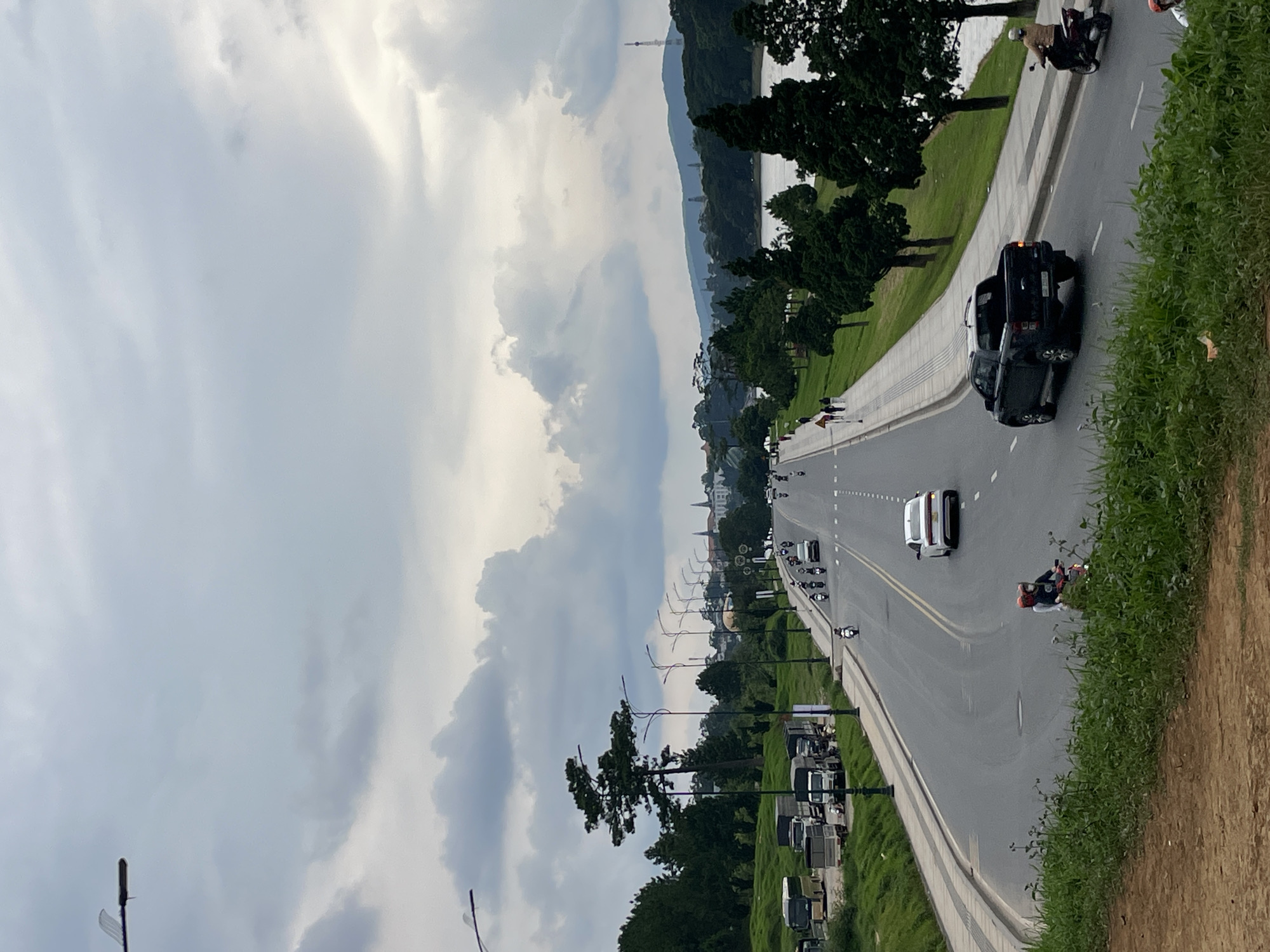 The image depicts a wide, curving asphalt road in Lâm Viên - Đà Lạt, Vietnam. The road is lined with a paved pedestrian walkway and grassy slopes dotted with cypress trees. On the left side of the road, parked trucks and buses are visible on a grassy incline. Traffic on the road includes several motorcycles, one white sedan, and a black pickup truck. Pedestrians are seen walking on the side of the road. In the background, a body of water is visible to the right, with rolling hills and distant buildings, including a prominent tower, forming the horizon under a cloudy sky. The overall impression is an urban landscape with natural elements, under a subdued, overcast sky.