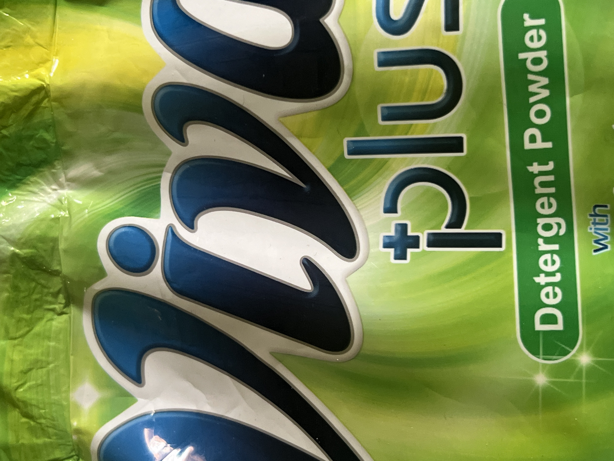 The image shows a close-up of a green and yellow package of "Viva Plus Detergent Powder." The brand name "Viva" is written in large, bold, blue letters with white outlines, dominating the upper portion of the image. Below it, the word "plus" is written in a smaller, white font with blue outlines, with a blue plus sign preceding it. The bottom part of the image contains the text "Detergent Powder" inside a green oval with a white outline. The packaging has a wrinkled, glossy texture. The background is a gradient of green and yellow, with subtle wavy lines. There are no people or any contextual environmental details visible that would relate to the city of Jauro Yinu, Nigeria.