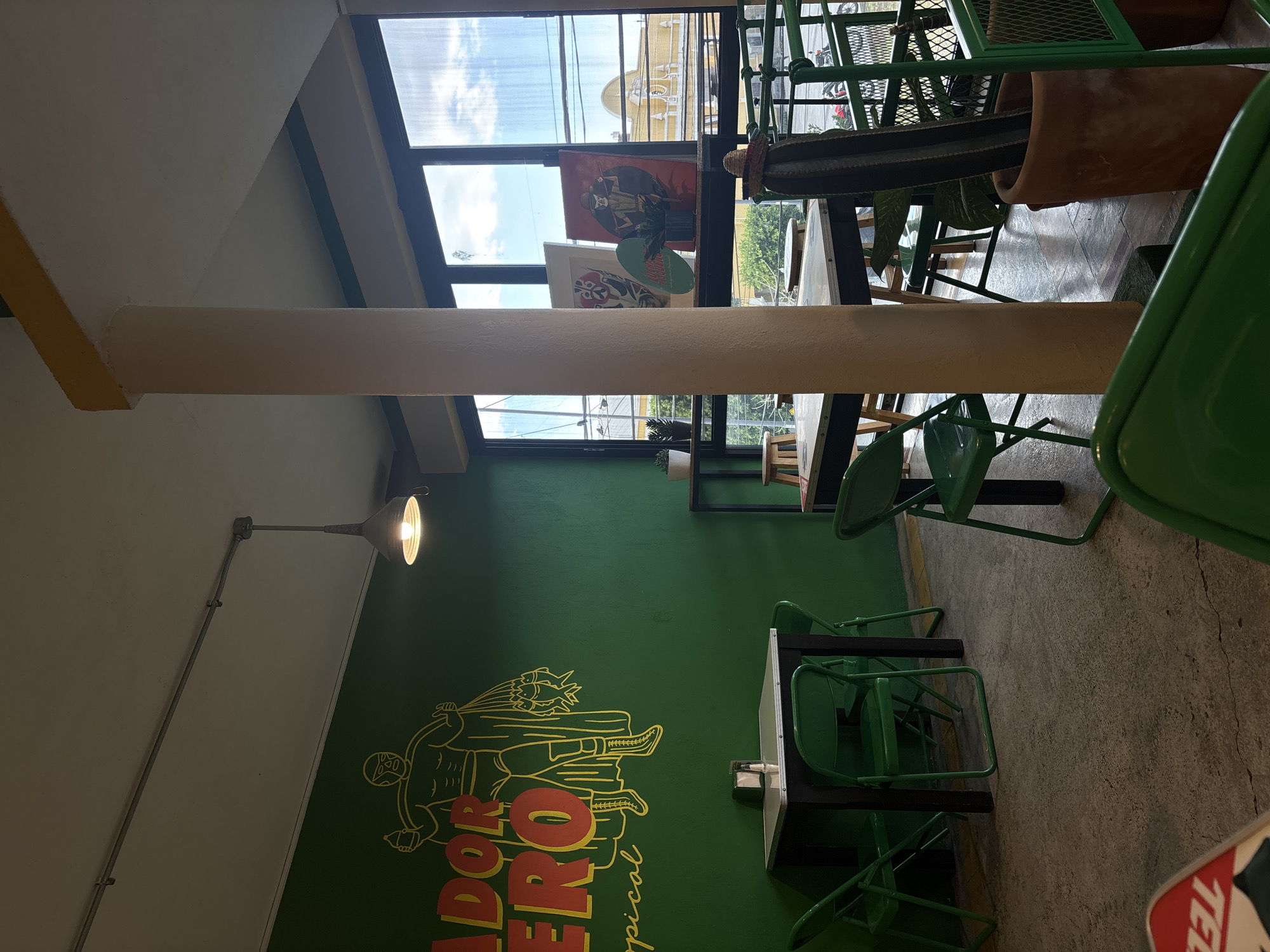 The image displays the interior of a casual dining establishment in San Andrés Cholula, Mexico. The left wall is painted bright green, featuring a large yellow-outlined mural of a lucha libre wrestler holding a bottle in one hand and a net containing fish in the other, with partial text reading "ADOR HERO tropical" visible below. The ceiling is white, with an exposed conduit pipe leading to a single industrial-style pendant light fixture. The flooring is rough concrete.
In the left-midground, a dark-framed table with a light-colored surface is equipped with a napkin dispenser and salt/pepper shakers, flanked by three bright green metal folding chairs. A tall, light-colored support pillar with a yellow capital section stands centrally, extending from the floor to the ceiling.
To the right of the pillar, large windows span the wall, offering a view of the exterior. The windows consist of multiple dark-framed panes, some appearing translucent or corrugated. Beyond the windows, a blue sky with scattered white clouds is visible, along with overhead power lines, green foliage, and a section of a red motorcycle or scooter on the street level. In the distance, a distinctive yellow domed building with decorative window patterns is identifiable.
A dark shelf runs along the base of the windows, displaying several framed artworks—including one with a red background and a figure wearing a hat—a round teal-colored sign, and small potted succulents. In the lower right-midground, a tall columnar cactus is positioned in a terracotta pot, with a miniature sombrero resting atop it. Parts of additional dark-framed tables with light tops, green metal folding chairs, and wooden stools are visible in the right section of the room. A green metal barrier or shelving unit is partially visible in the lower right foreground. No individuals are present in the scene.