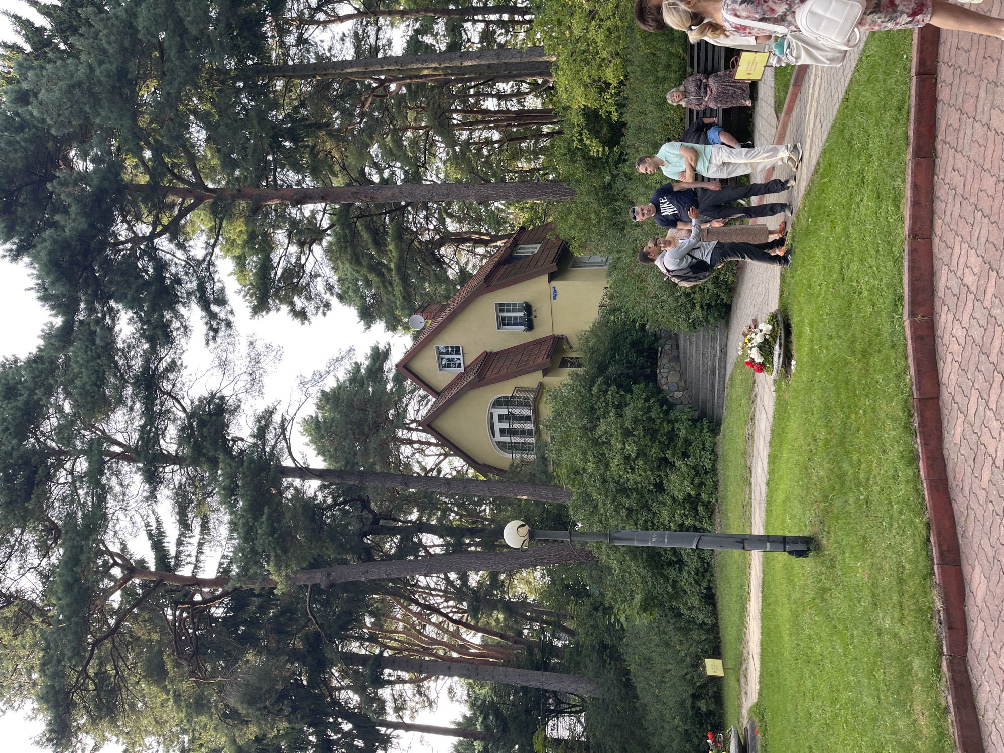 A group of people are gathered in a park setting in front of a yellow house with a red roof. The house is two stories tall with several windows and a balcony. It is surrounded by tall pine trees and lush green bushes. In the foreground, there is a paved walkway and a grassy area with a flower planter. Several people are standing and talking, while others are sitting on a bench. One person is walking away from the camera, carrying a white bag. The lighting suggests it is daytime, with sunlight filtering through the trees. The scene appears to be in the city of Svetlogorsk, Russia, as indicated by the architectural style and the presence of mature pine trees commonly found in the region.