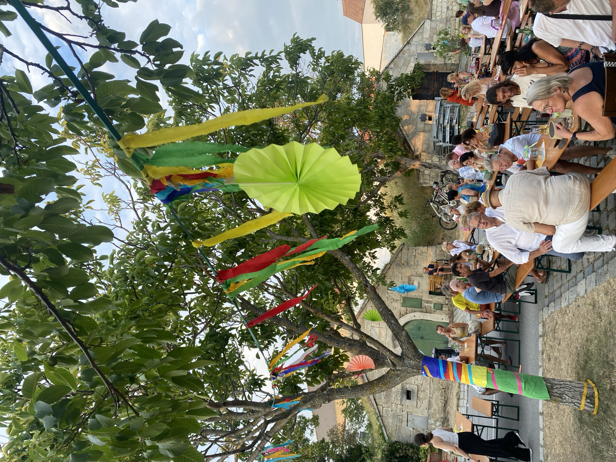 The image depicts an outdoor gathering at a wine tavern in Purbach, Austria, likely during the evening. The scene is set beneath a large tree adorned with colorful streamers and paper decorations. Several long wooden tables are set up, with people seated at them, engaged in conversation and dining. Some individuals are standing, while others are seated on benches. The background features a rustic stone building with a green door and a green grass roof, characteristic of the Burgenland region. The overall atmosphere suggests a festive social event.