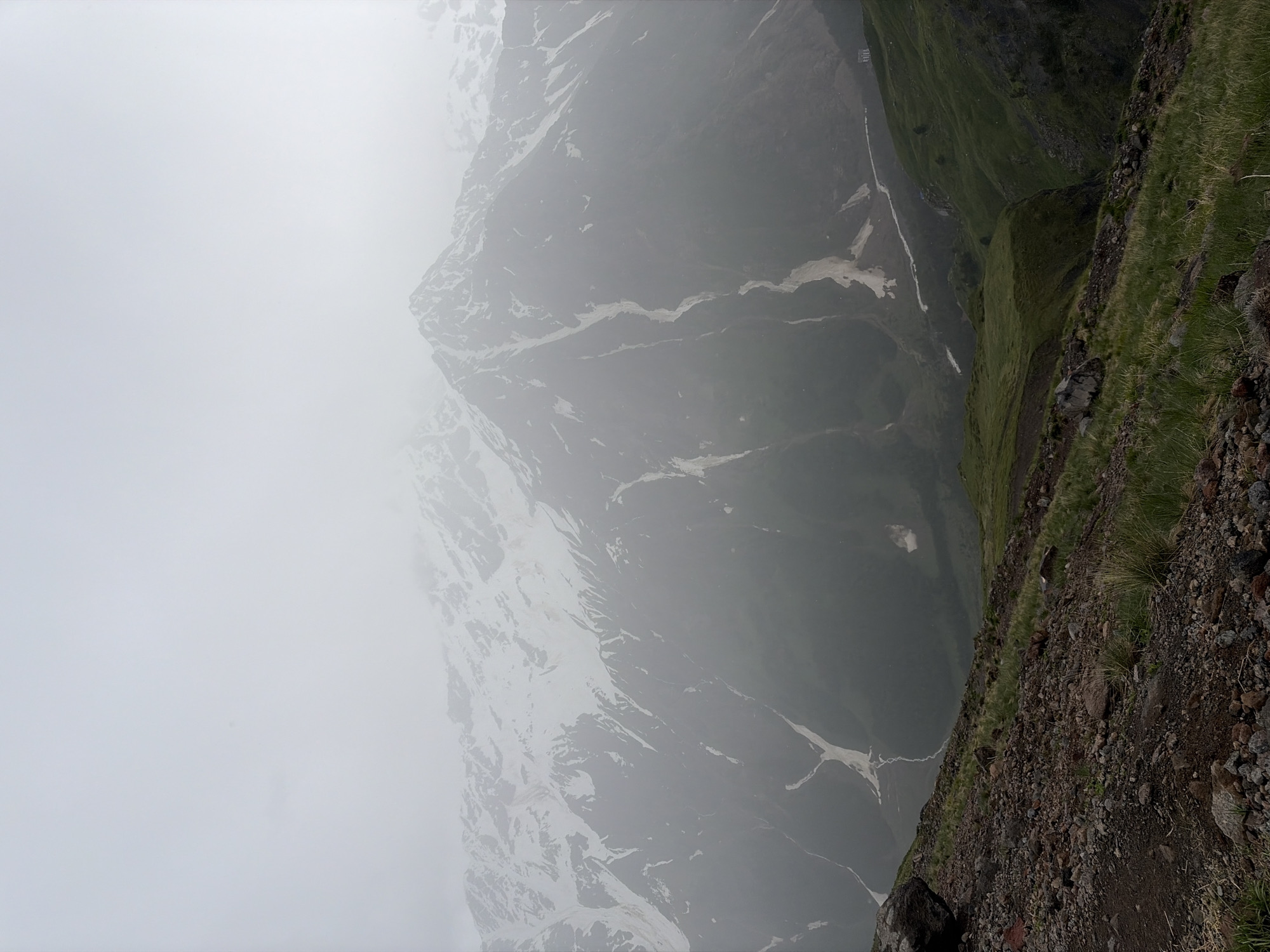 This image captures a view of Mount Elbrus, the highest peak in the Caucasus Mountains, near the town of Terskol, Russia. The scene is shrouded in fog, obscuring much of the mountain's upper reaches and the sky. The foreground is a rocky, barren slope with sparse green grass.
In the midground, the lower slopes of the mountain are covered in green vegetation and scree. Patches of snow are visible on the mountainside, interspersed with dark rock and earth. A winding path or a dried riverbed can be seen cutting through the landscape, leading towards the base of the mountain. In the distance, a small, white building is perched on a green hillside, adding a human element to the vast natural landscape. The overall atmosphere is one of mist and elevation, highlighting the rugged beauty of the mountainous terrain.